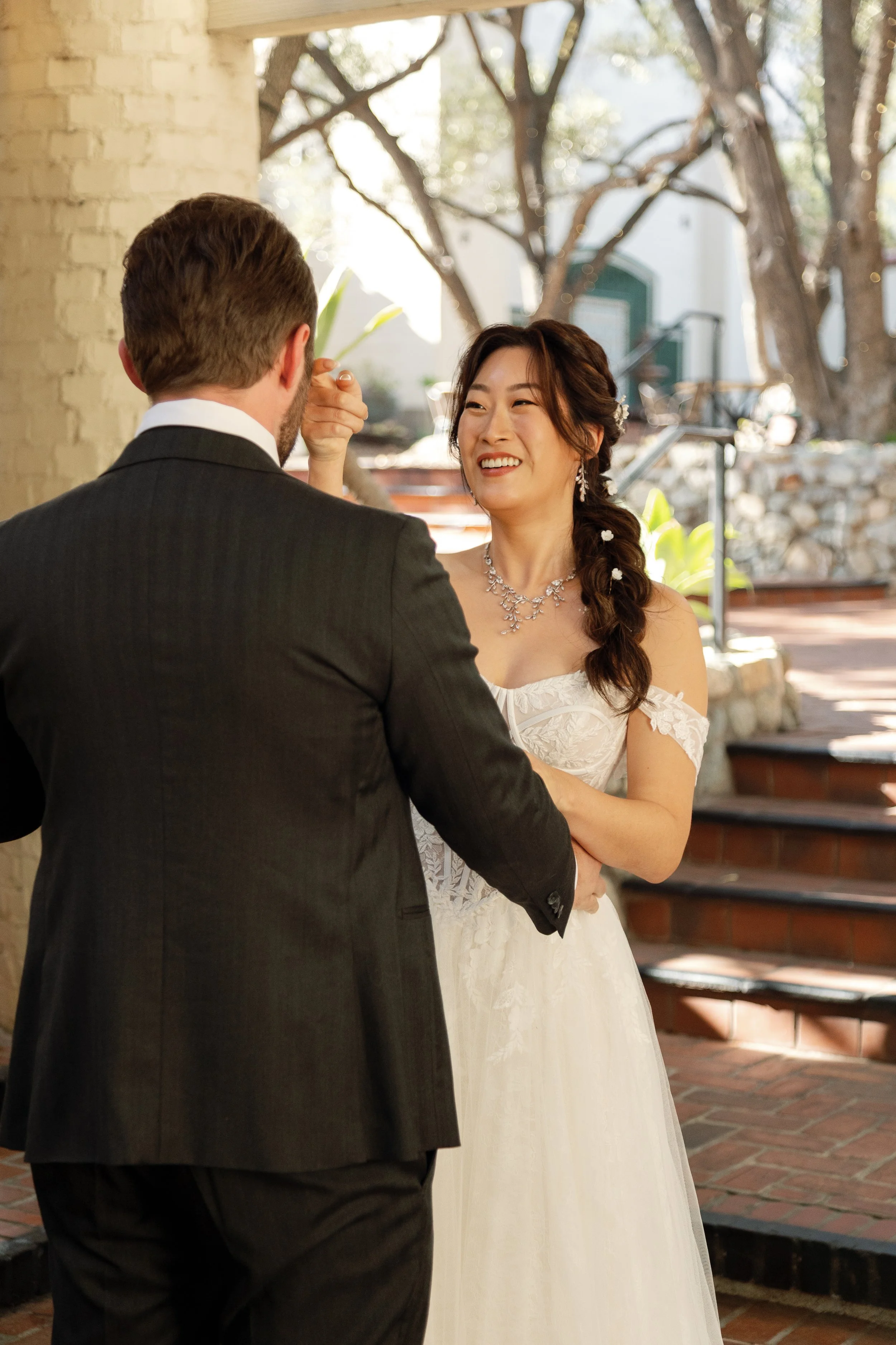 A bride and groom with their backs to the camera, sharing a moment during their wedding's first look outdoors on a sunny day, with steps and trees in the background.