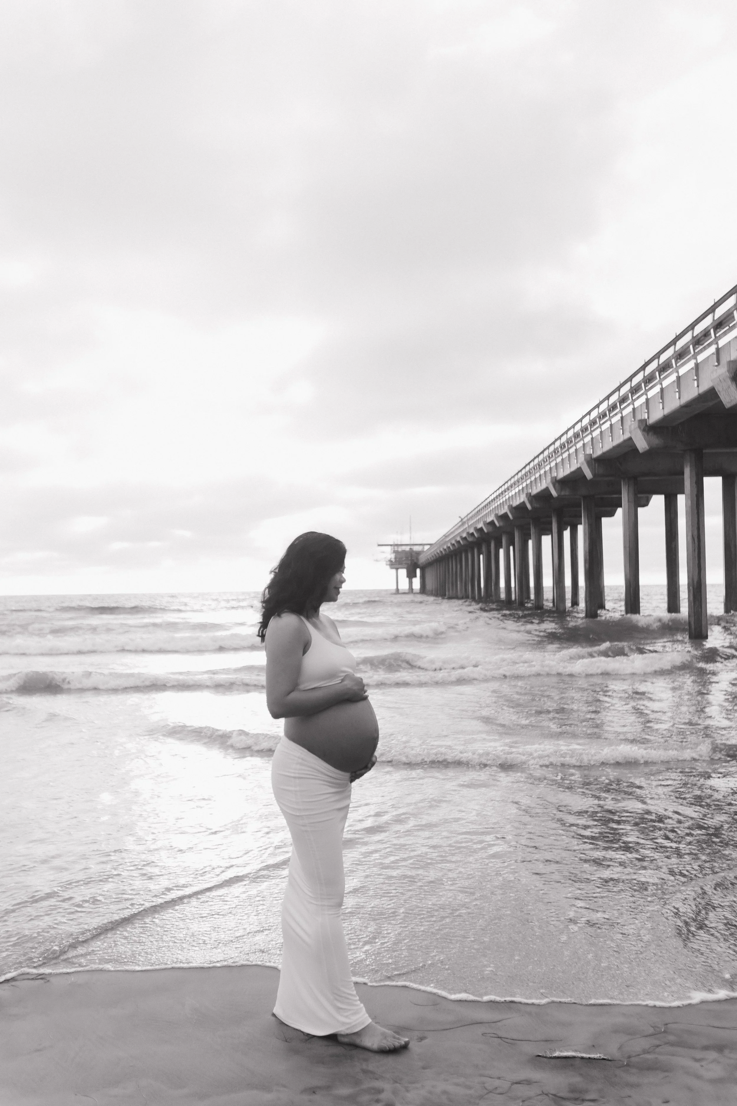 A pregnant woman standing barefoot on the beach near the ocean, gently holding her belly, with a pier extending into the water in the background, in black and white during Maternity photoshoot.