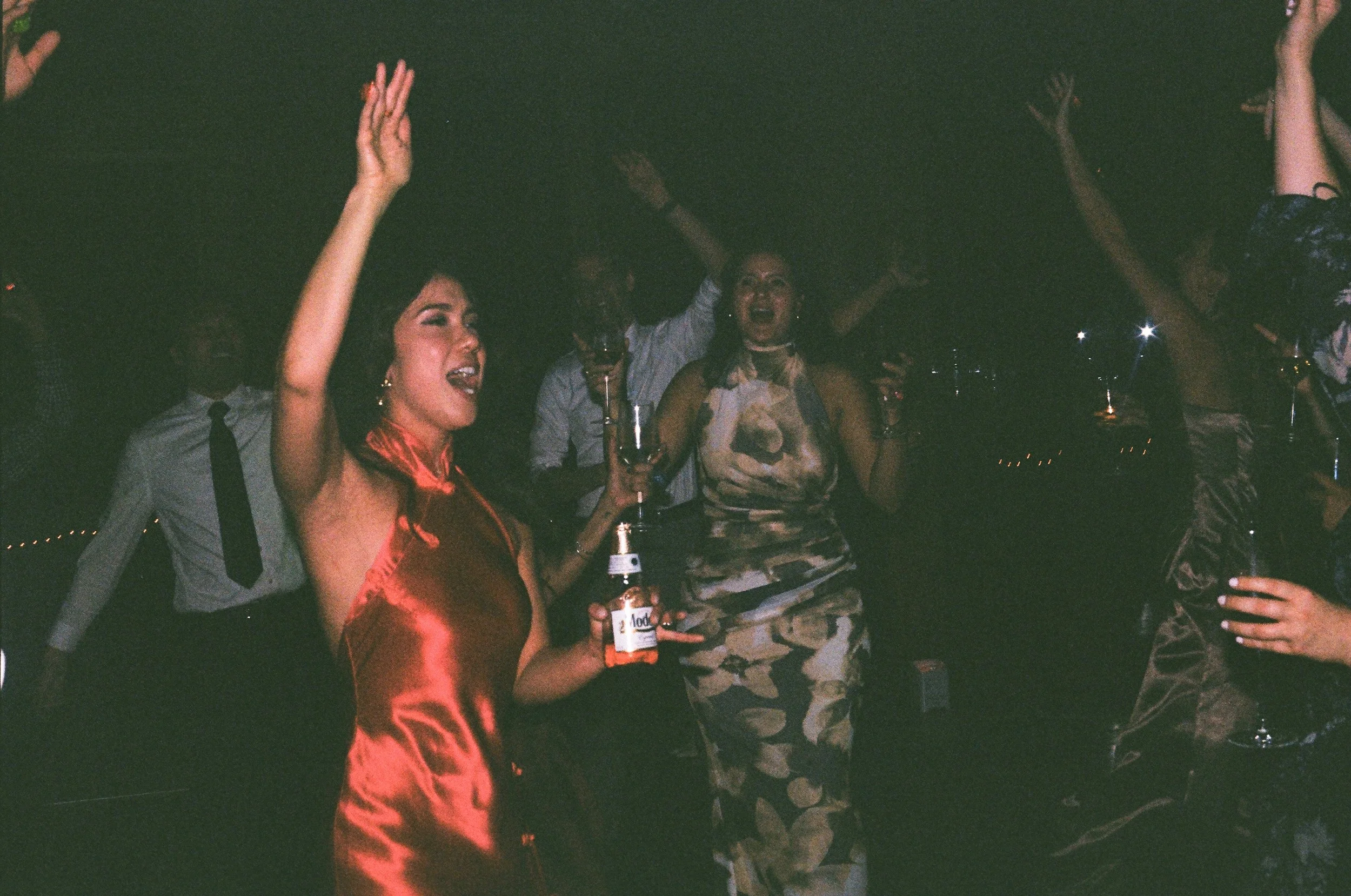 People dancing and celebrating at a wedding reception during the night, some holding drinks, with joyful expressions and raised arms.