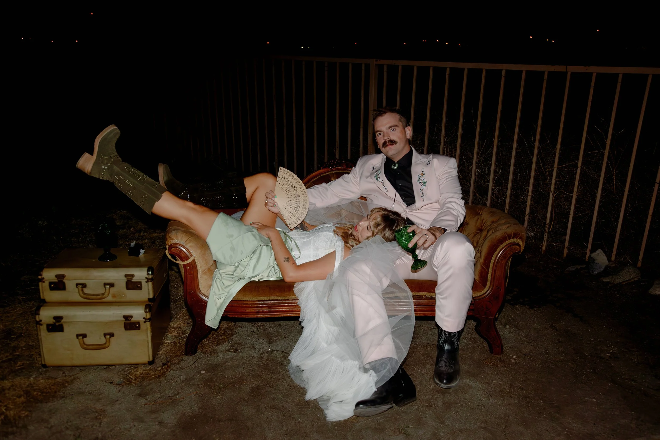 A man and woman lie on a vintage sofa outdoors at night, surrounded by a black fence. The man wears a white suit with embroidery, holds a green drink, and looks at the camera. The woman is dressed in a white dress with a green skirt, holding a fan, a