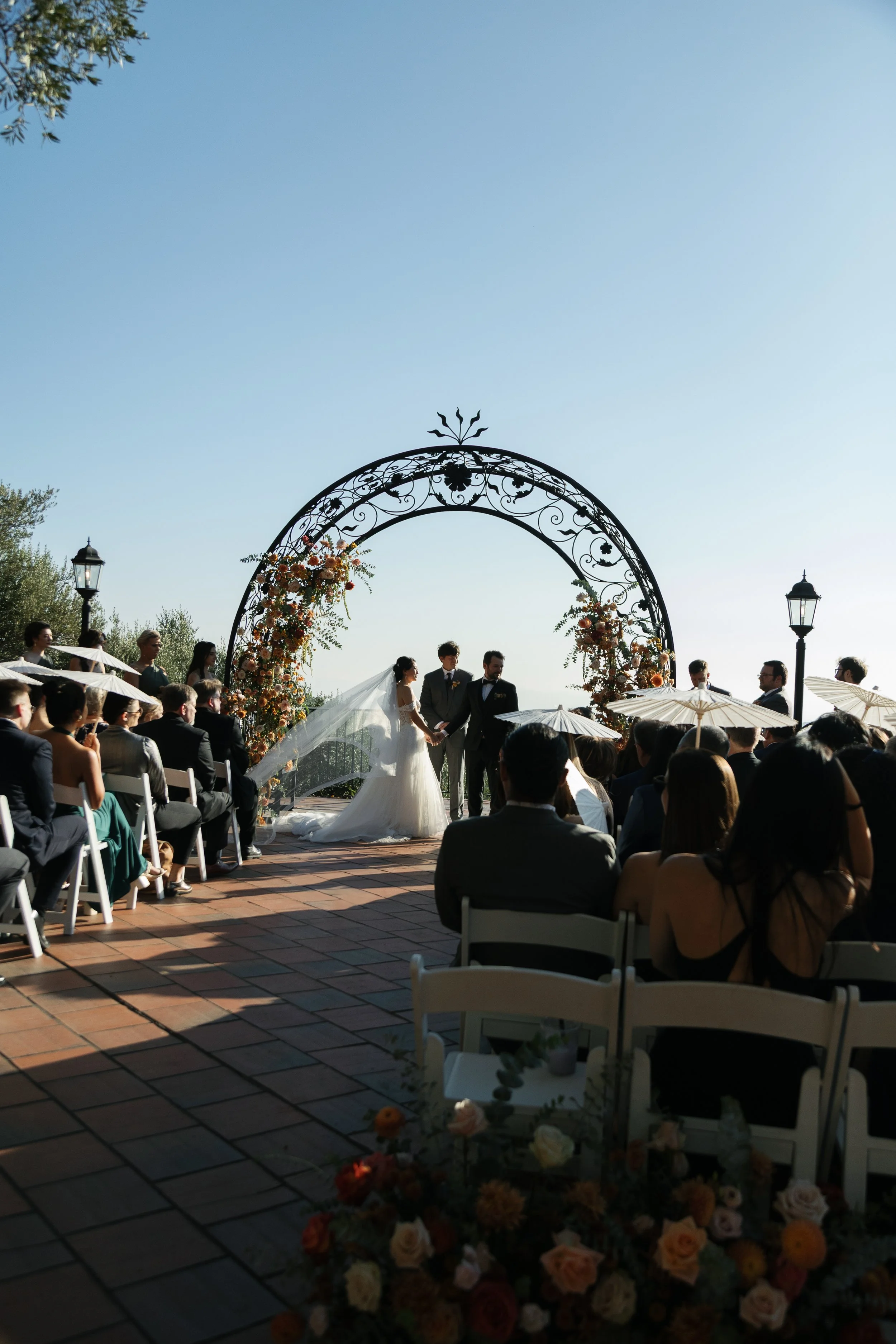 A wedding ceremony taking place outdoors under a metal arch decorated with flowers. The bride and groom are holding hands, with the officiant standing in front of them. Guests are seated on white chairs, some holding umbrellas for shade, surrounded by trees and lampposts.
