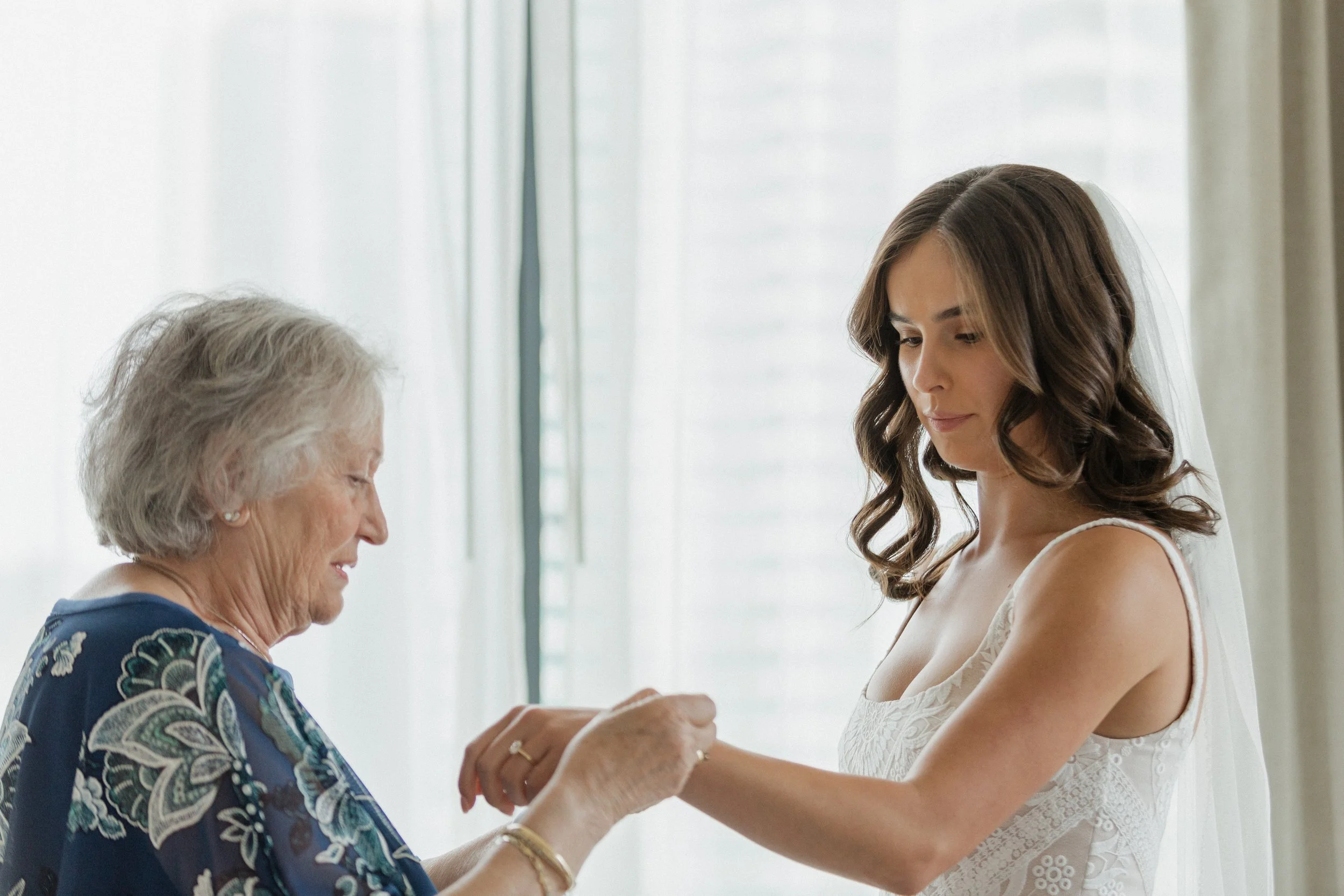 A young woman in a wedding dress, a bride, having her grandmother put on her bracelet, during getting ready photos indoors.