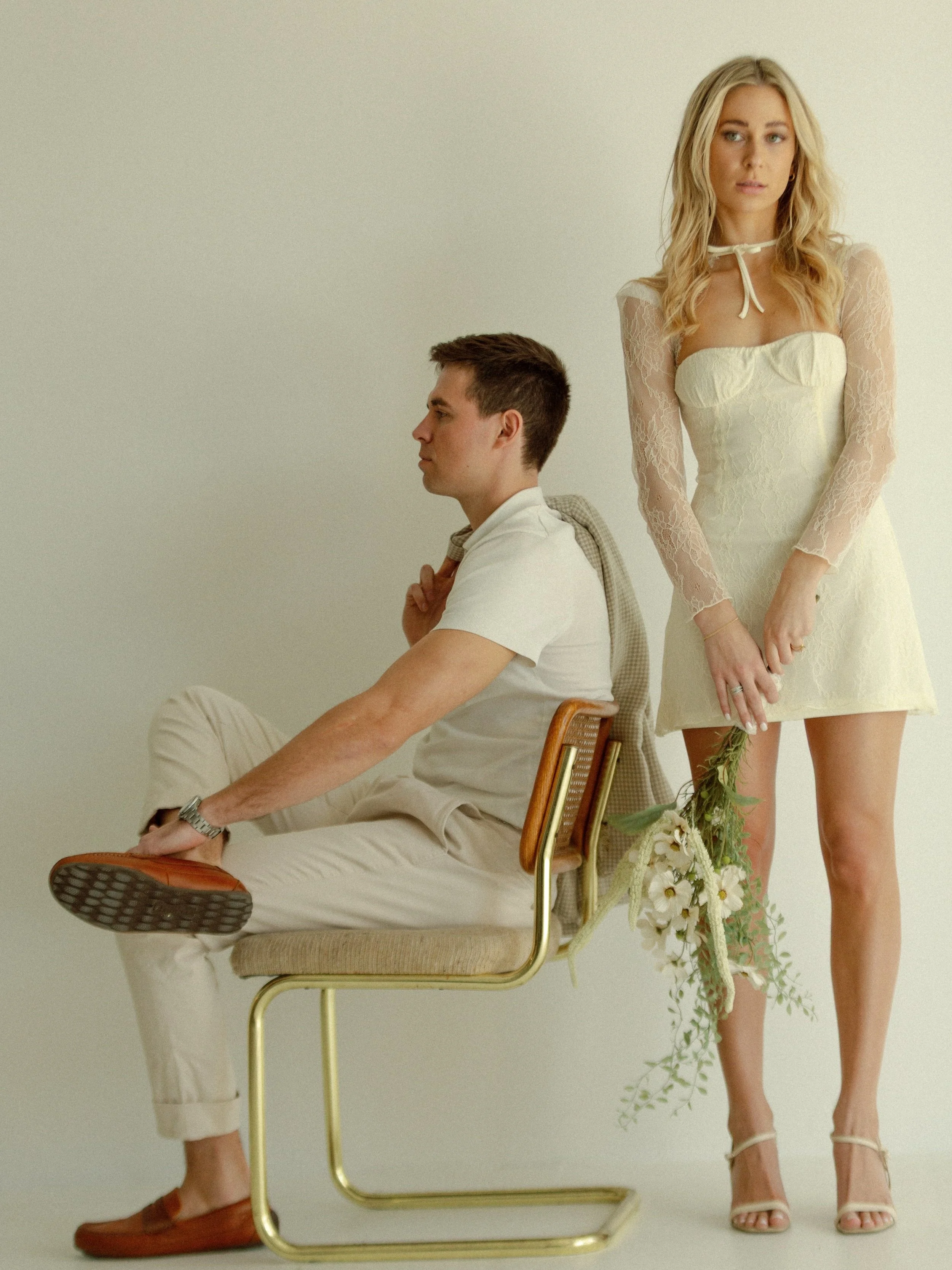 A man sitting on a chair with a woman standing behind him, holding a bouquet of flowers, in a minimalist setting with a neutral background. The woman is wearing a cream-colored dress with lace sleeves and high heels, while the man is dressed casually in a white shirt and beige pants.