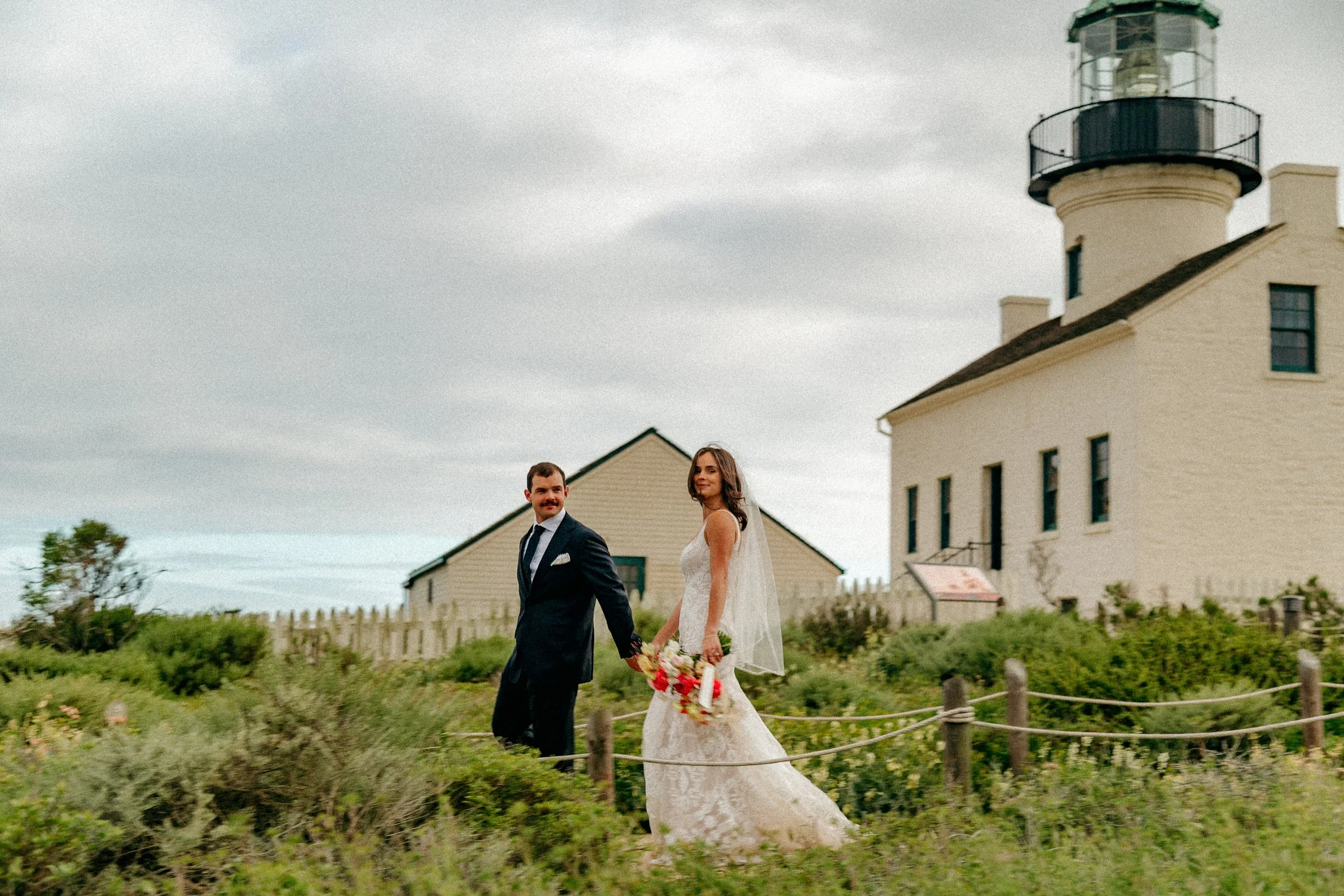 A bride and groom holding hands and walking outdoors near a lighthouse. The bride is in a lace wedding dress holding a bouquet, and the groom is in a dark suit. The lighthouse is tall with white walls and a green top, and there are cloudy skies overhead.