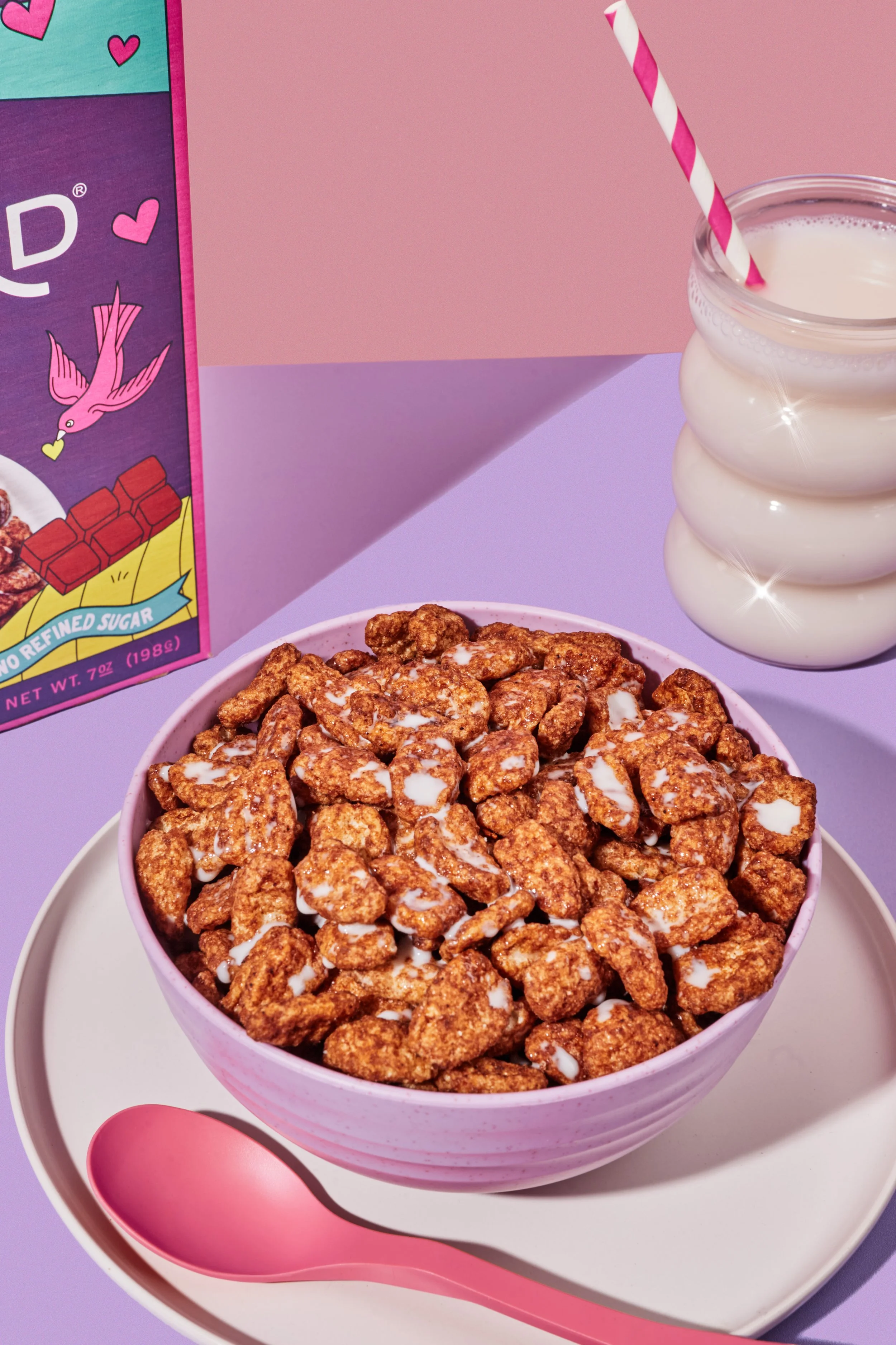 A pink bowl filled with chocolate cereal with white milk, placed on a off-white plate with a pink spoon, next to a glass of milk on a pastel purple and pink background. Part of a colorful cereal box is visible on the left.
