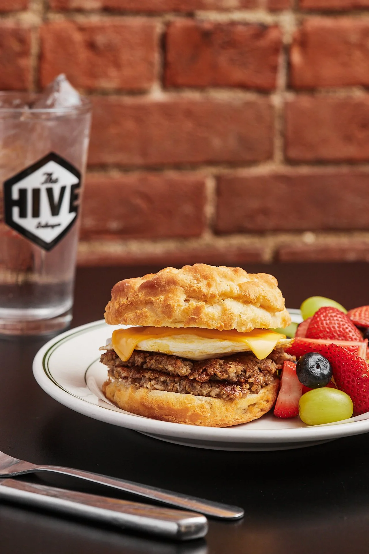Plate with sausage, fried egg, and biscuit sandwich alongside mixed fruit, with a glass of water in the background against a brick wall