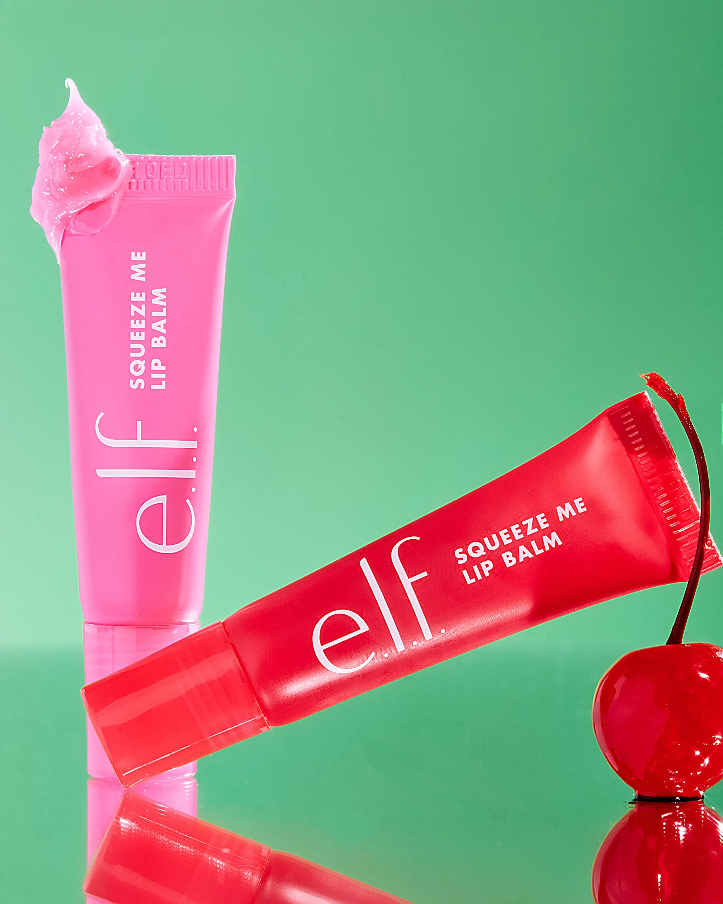 Two tubes of elf Squeeze Me Lip Balm, one pink and one red, with a cherry beside them, on a green background.