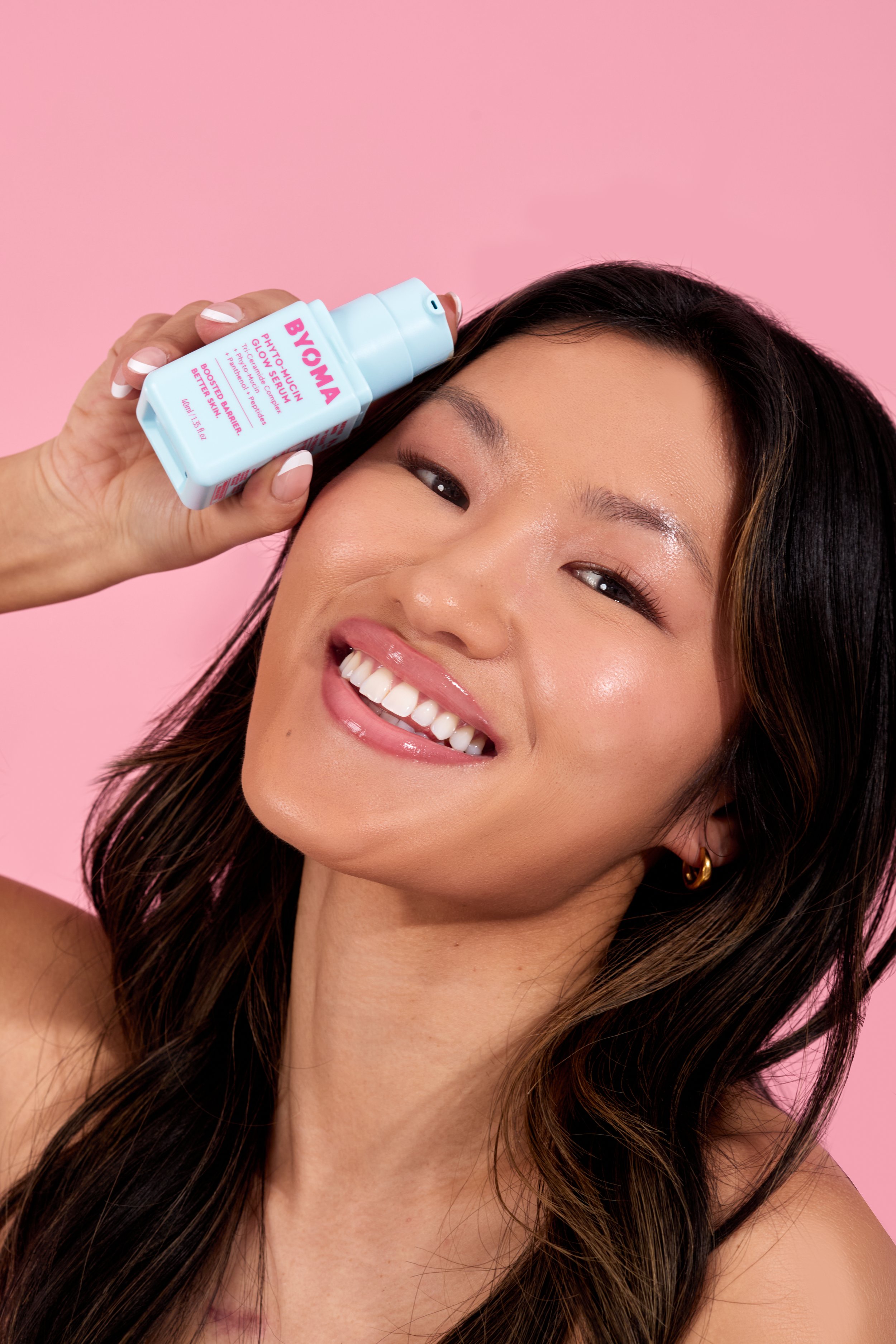 A smiling woman with long dark hair holding a skincare spray labeled 'BYOMA' near her temple against a pink background.