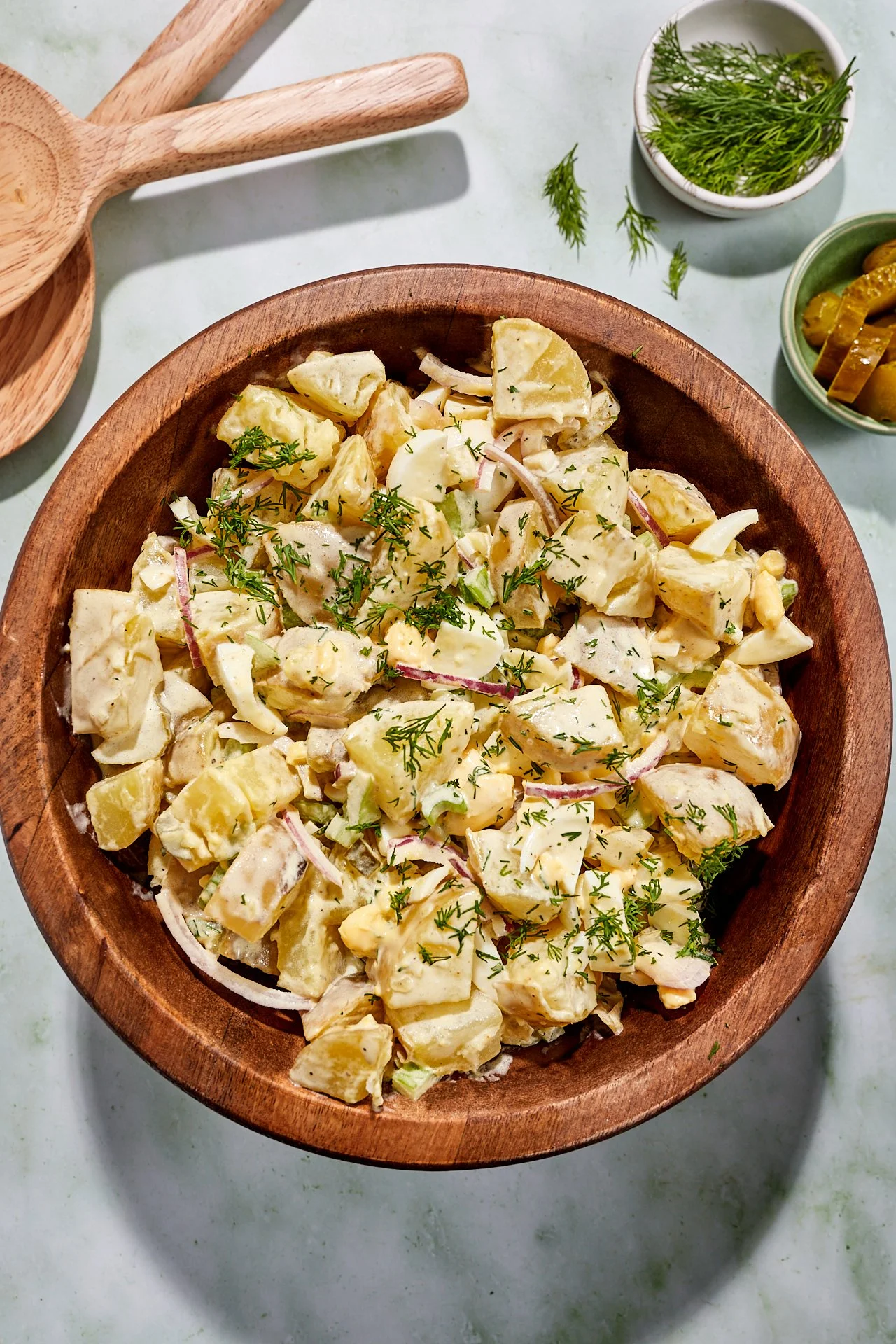 Potato salad garnished with fresh herbs in a wooden bowl, with bowls of fresh herbs and pickles nearby.