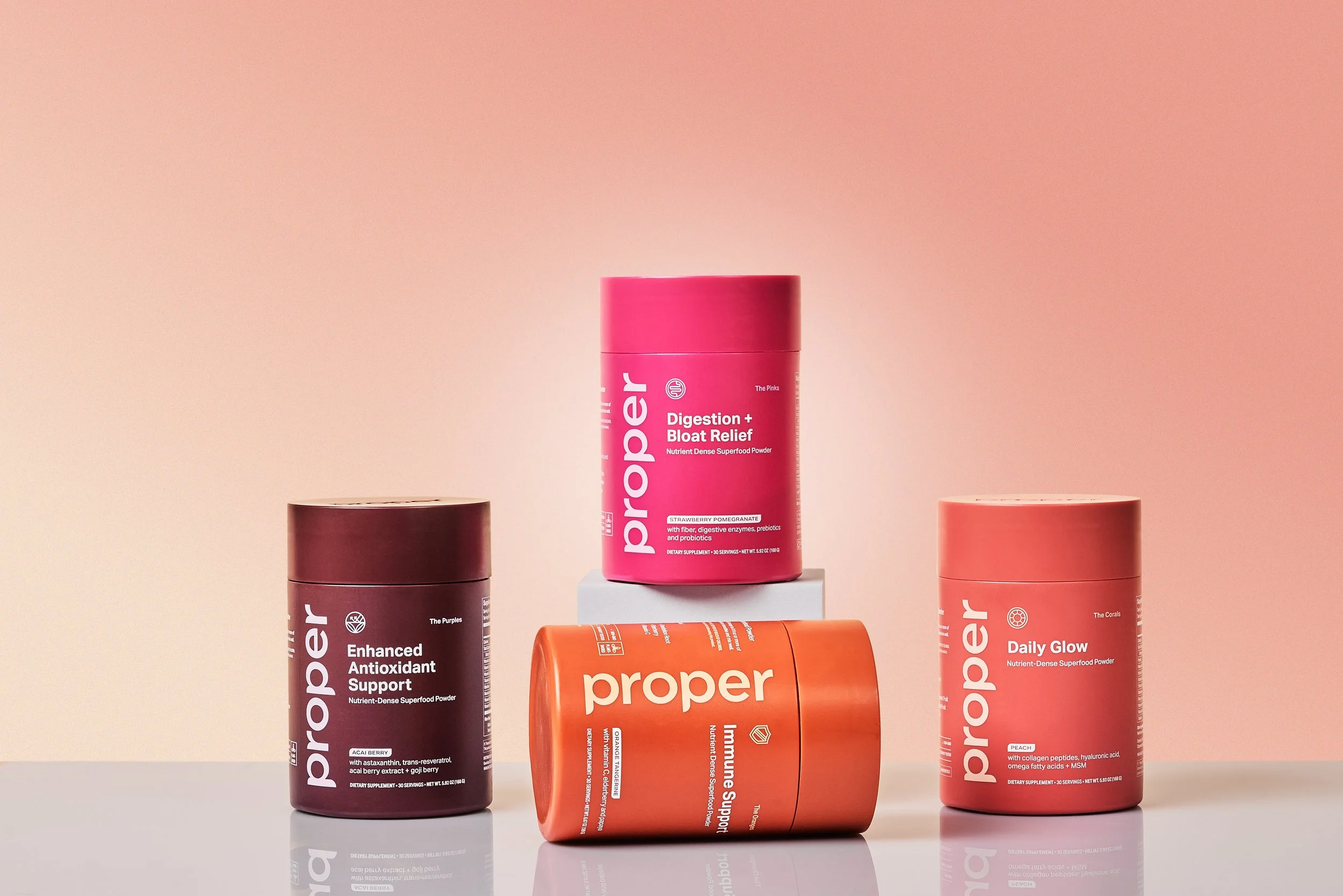 Collection of four colorful supplement containers from Proper, including Digestion + Bloat Relief, Enhanced Antioxidant Support, Daily Glow, and Immune Support, arranged on a white surface against a pink gradient background.