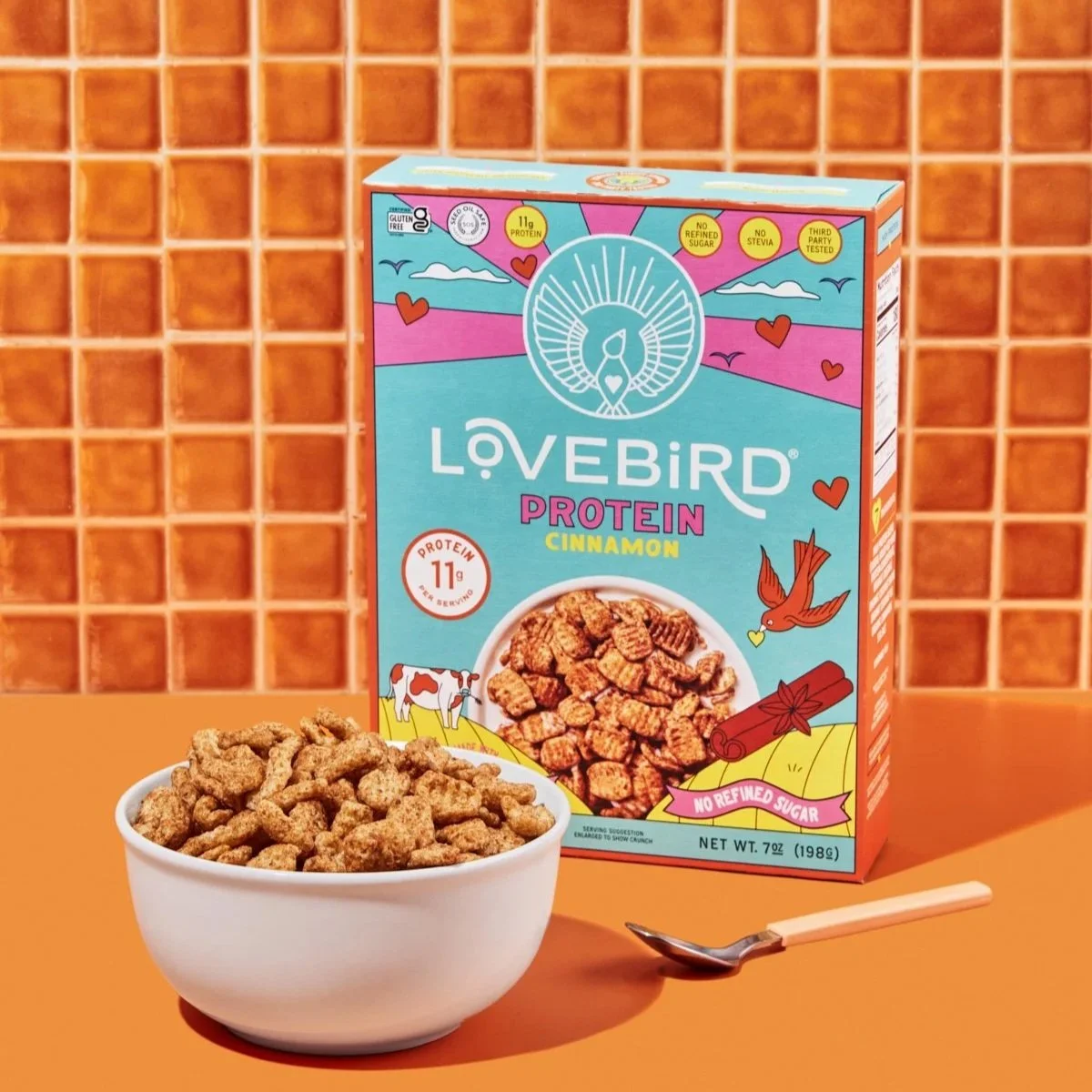 A box of Lovebird Protein Cinnamon cereal, a white bowl filled with cereal pieces, and a spoon on an orange surface with a matching orange tiled wall in the background.
