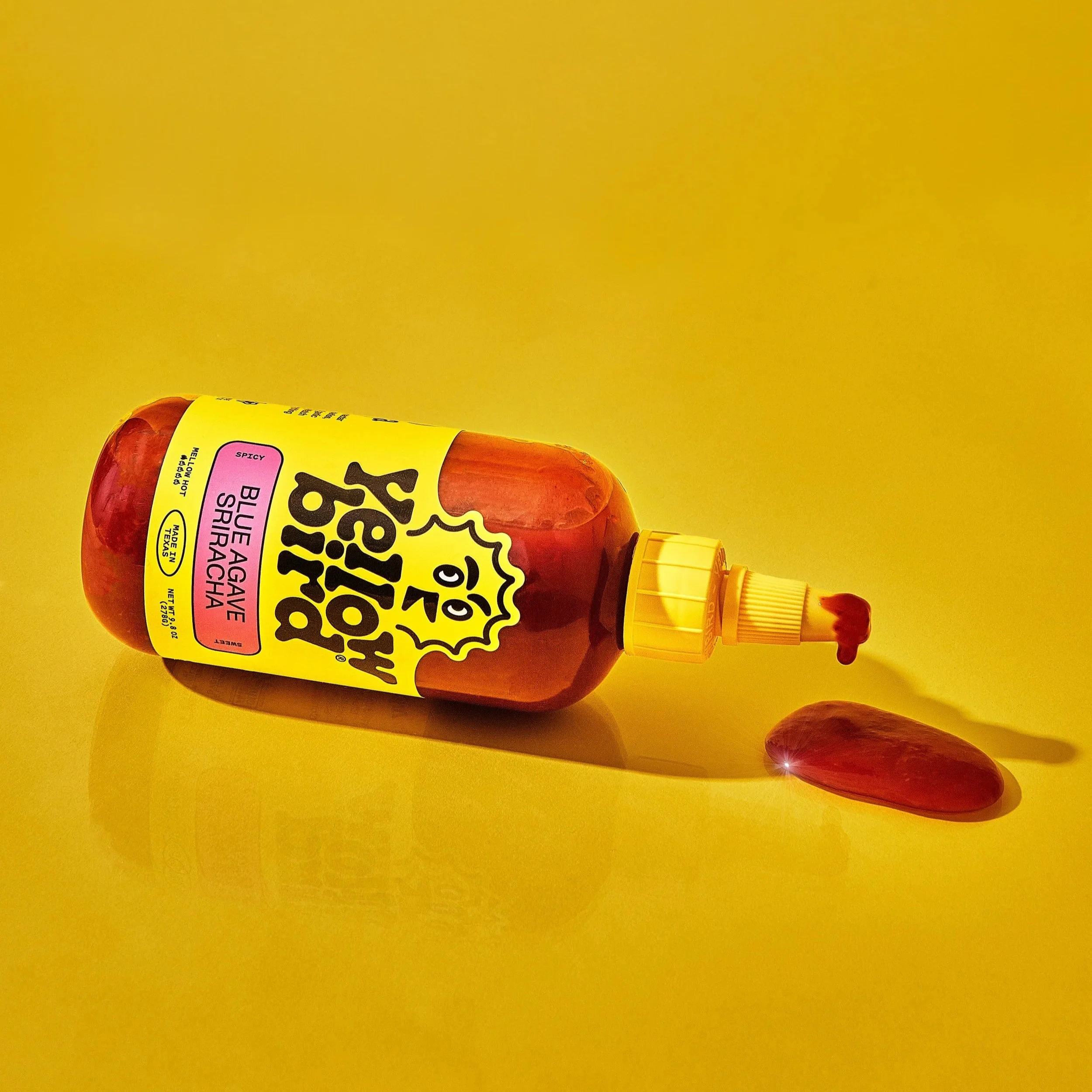 A bottle of Big Top Blue Aggave Sriracha hot sauce lying on its side on a yellow background. The bottle has a yellow cap with a small amount of red sauce leaking out. A drop of sauce is on the surface next to it, with a bright reflection.