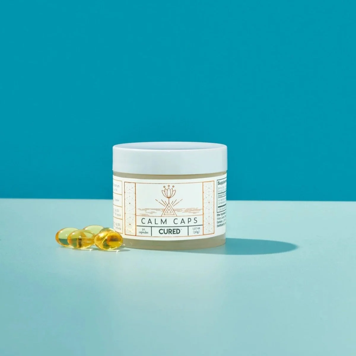 A white container labeled 'Calm Caps Cured' with 30 capsules, placed on a blue surface with a blue background. Four yellow capsules are beside the container.