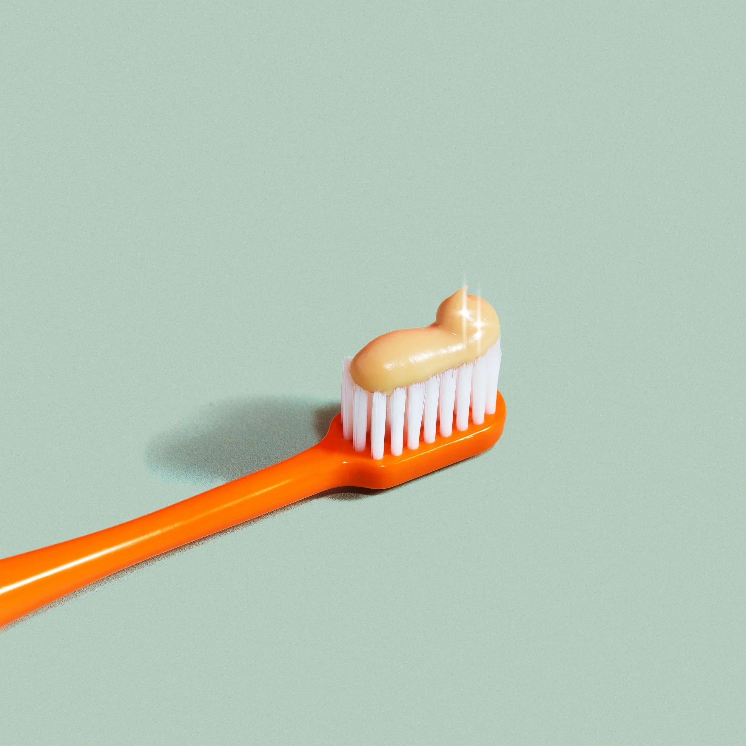 Toothbrush with orange handle and white bristles, with a dollop of yellow toothpaste on it, against a light green background.