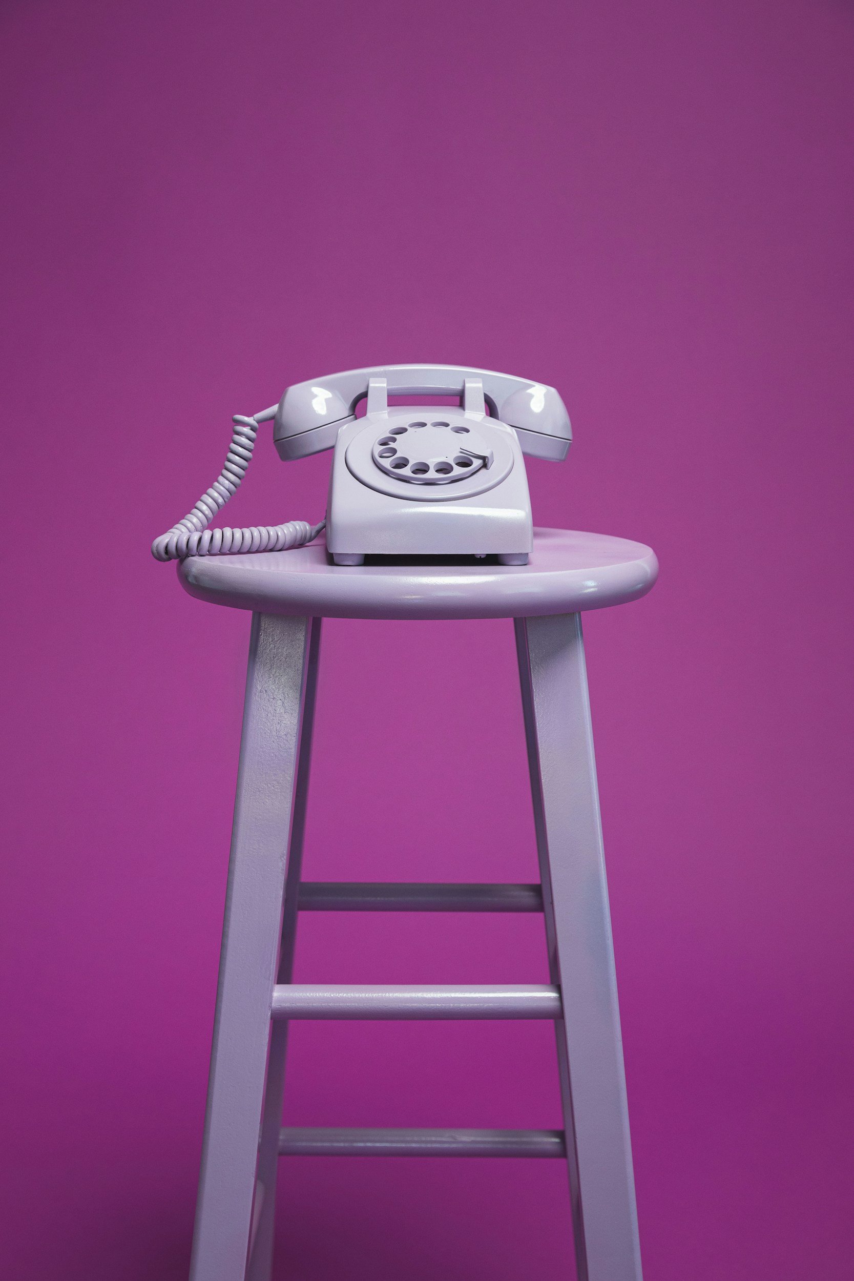 A retro rotary telephone in white, placed on a purple stool against a solid purple background.