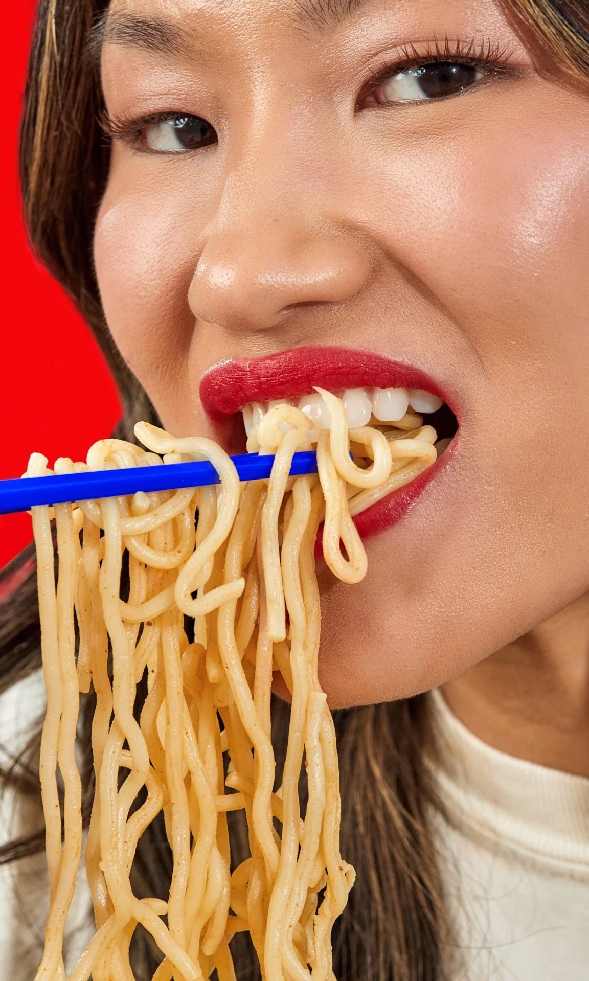 A woman with red lipstick eating noodles with blue chopsticks against a red background.