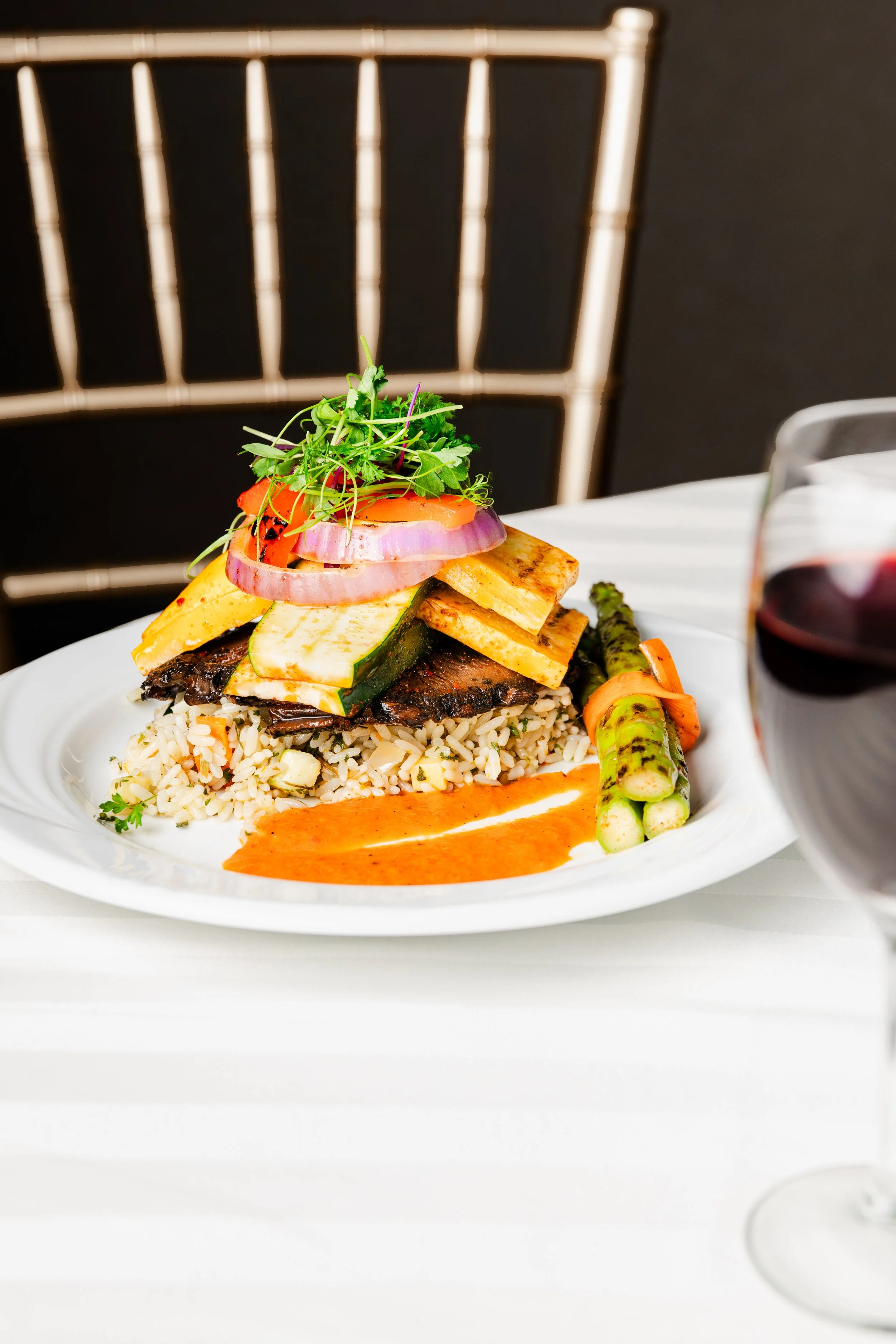 A plated dish with rice, grilled vegetables, grilled tofu, and a sauce, served with asparagus and a glass of red wine.