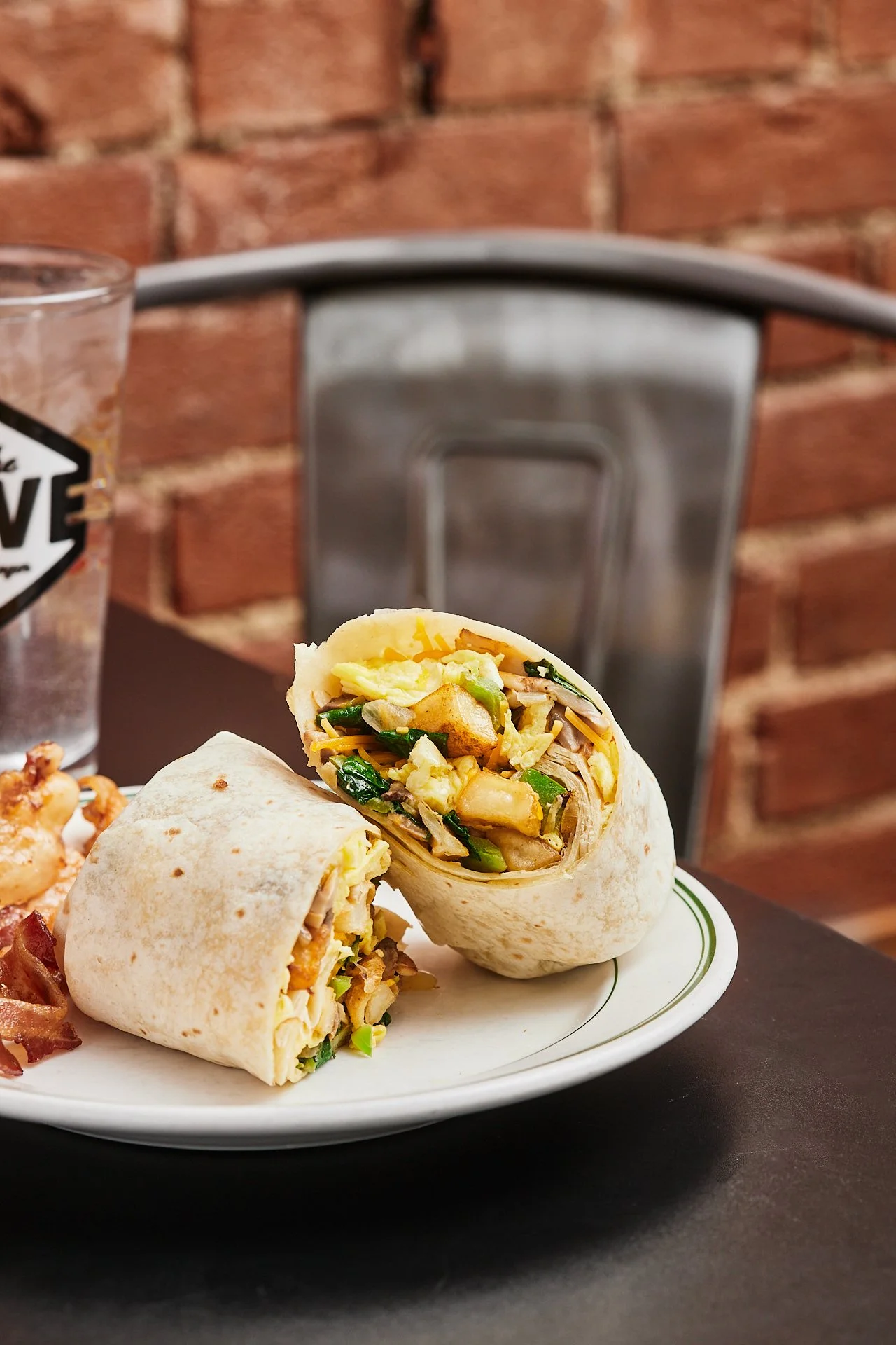 Close-up of a sliced burrito filled with vegetables and chicken on a white plate, placed on a black table in a restaurant with brick wall background.