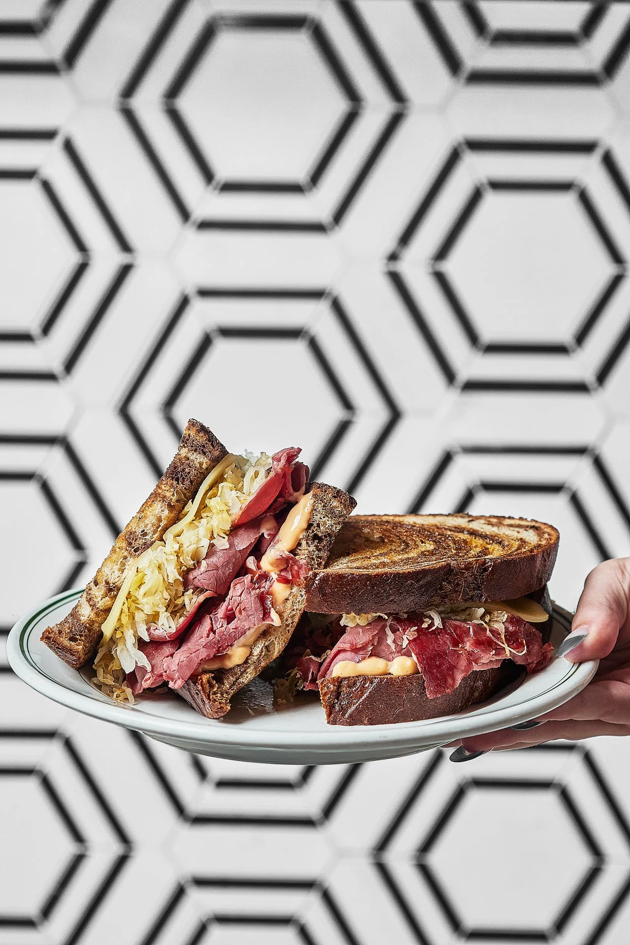 A hand holding a plate with two Reuben sandwiches, showing rye bread, corned beef, sauerkraut, cheese, and Russian dressing.