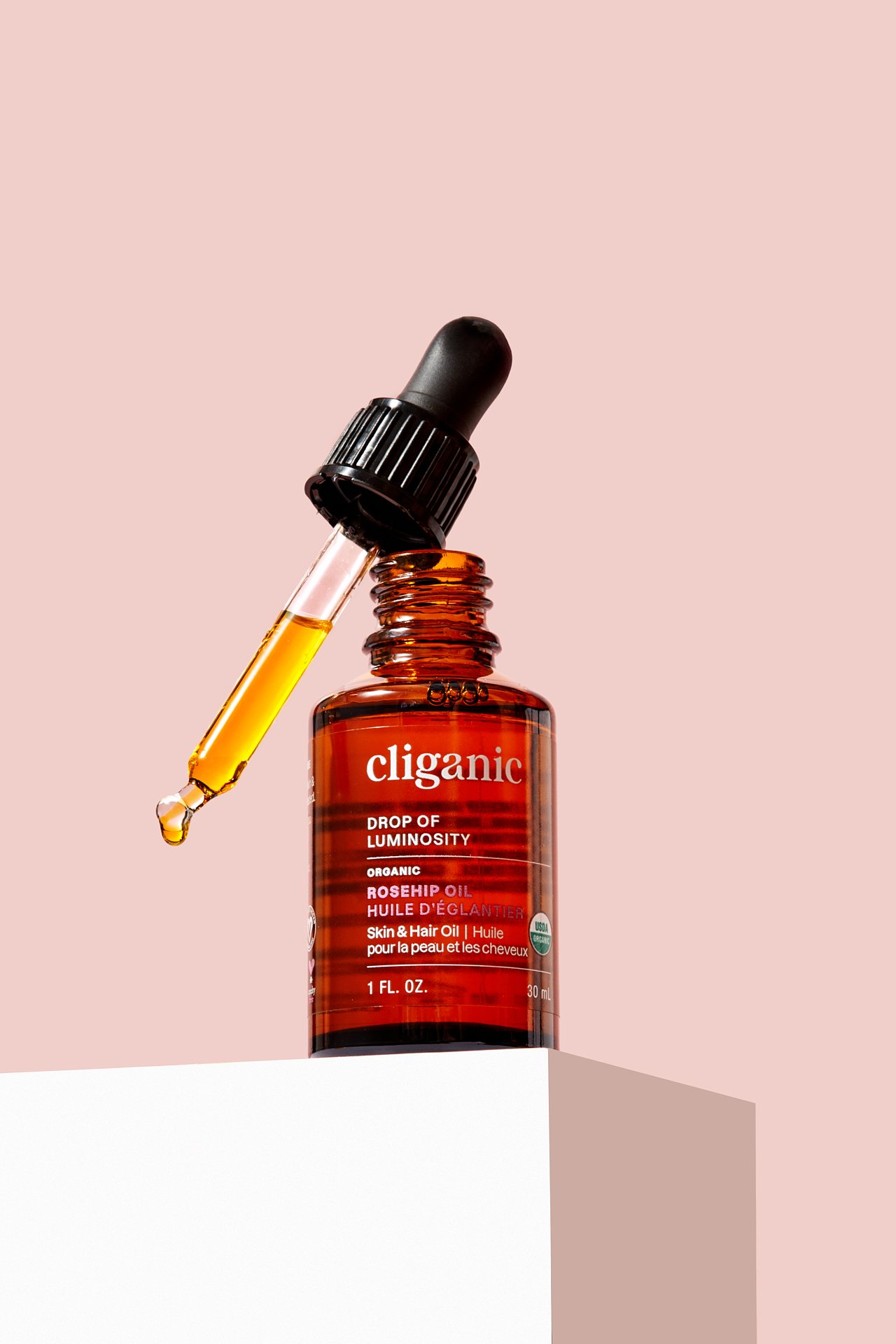 A small amber glass bottle labeled "cliganic" with a black dropper cap, and the dropper filled with a yellowish oil, set against a pink background.