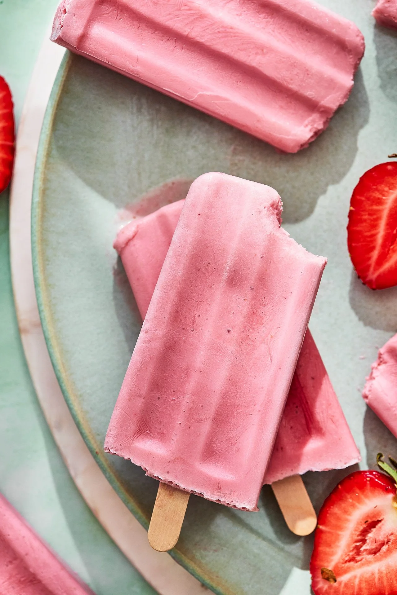 Two pink strawberry-flavored popsicles on a light-colored plate, with sliced strawberries nearby.