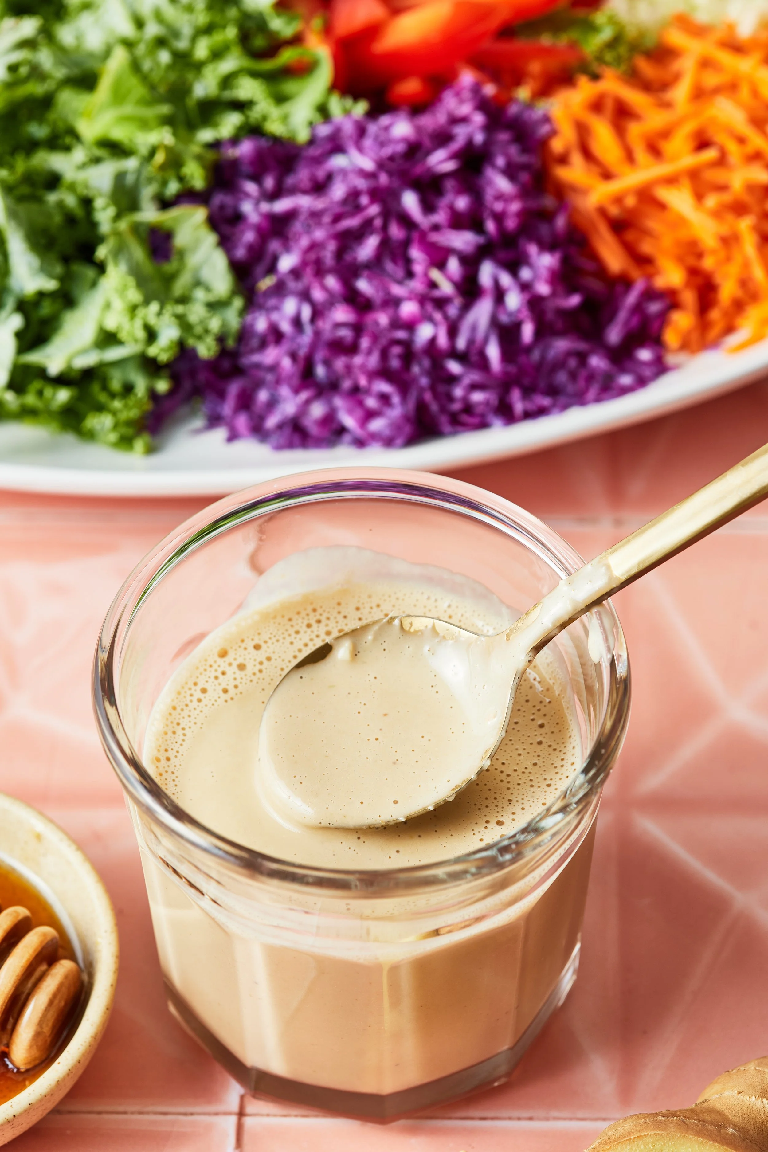 A glass of creamy dressing with a spoon inside, placed on a pink tiled surface, with a white plate of assorted shredded vegetables including purple cabbage, orange carrots, and green lettuce in the background.