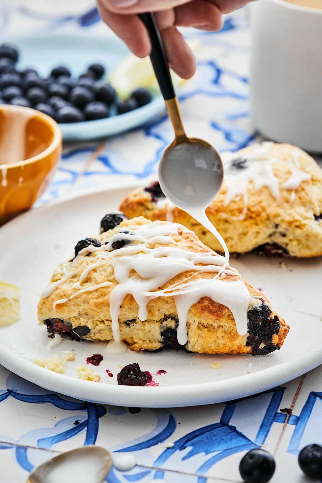 A slice of blueberry scone being drizzled with white icing on a white plate with a whole scone in the background, blueberry and lemon slices nearby, and bowls of blueberries and a beverage in the background.