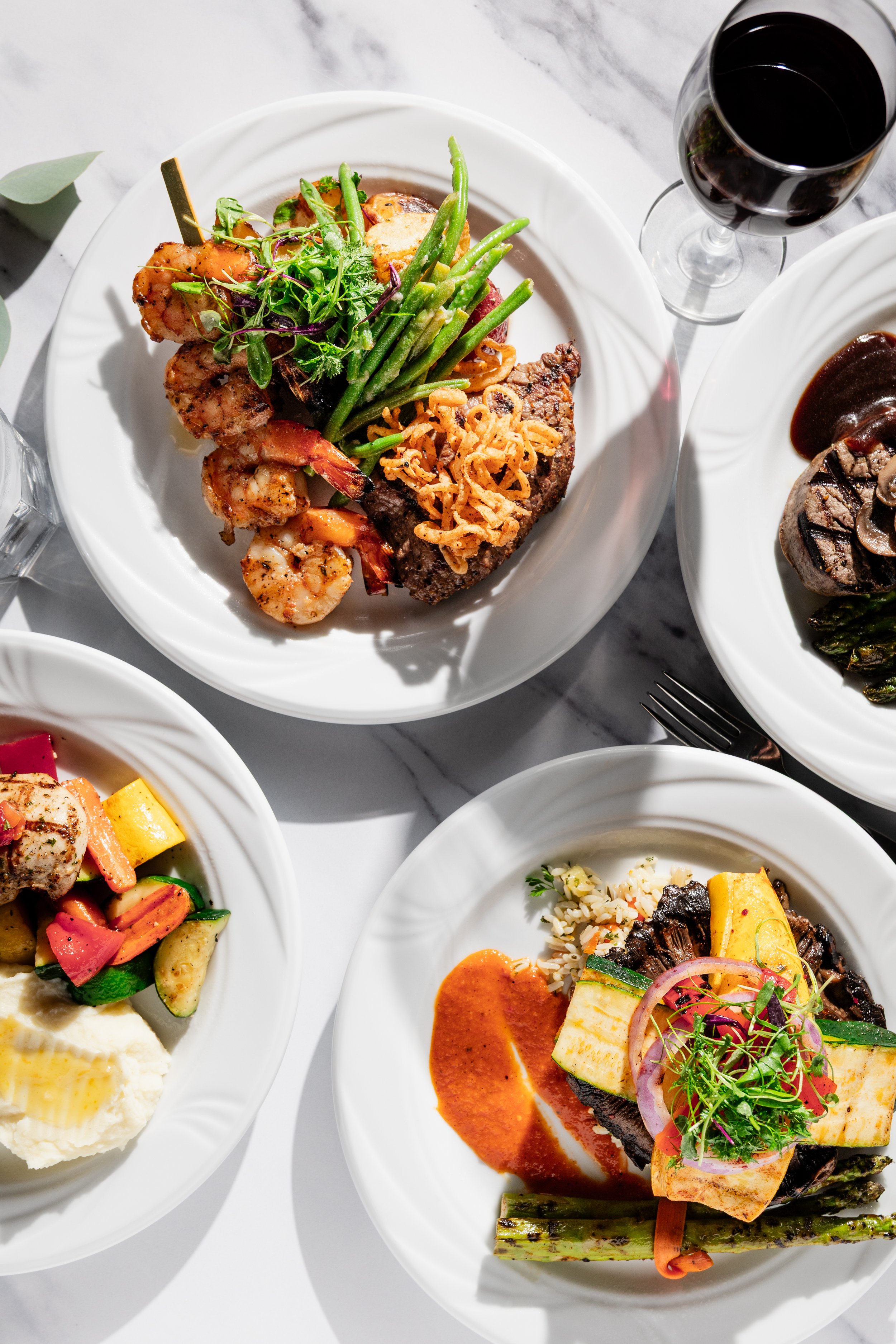Various plated gourmet dishes with seafood, vegetables, and meat, along with a glass of red wine on a marble table.
