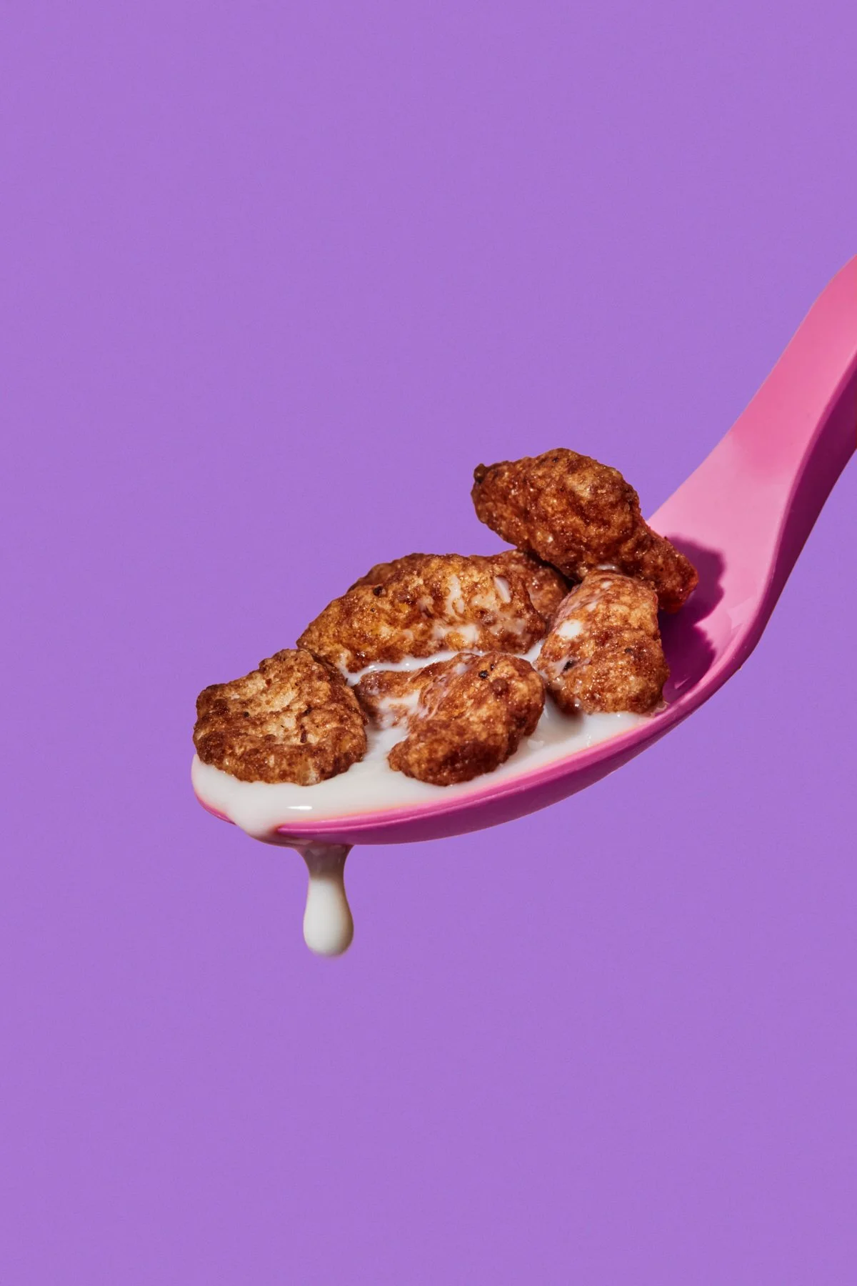 A pink spoon holding cereal pieces with milk, against a purple background.