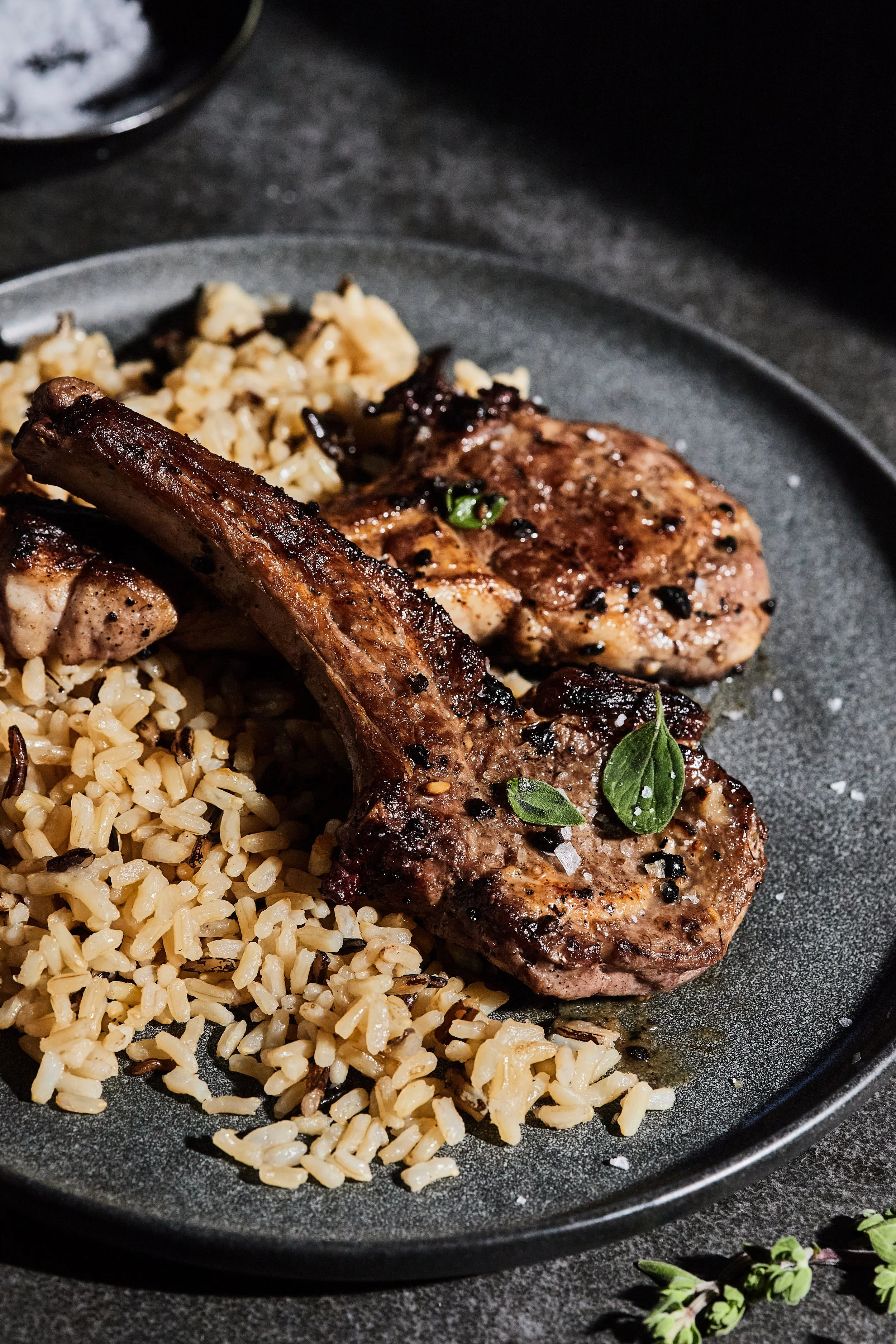 Grilled pork chops served with rice and garnished with fresh herbs on a black plate.