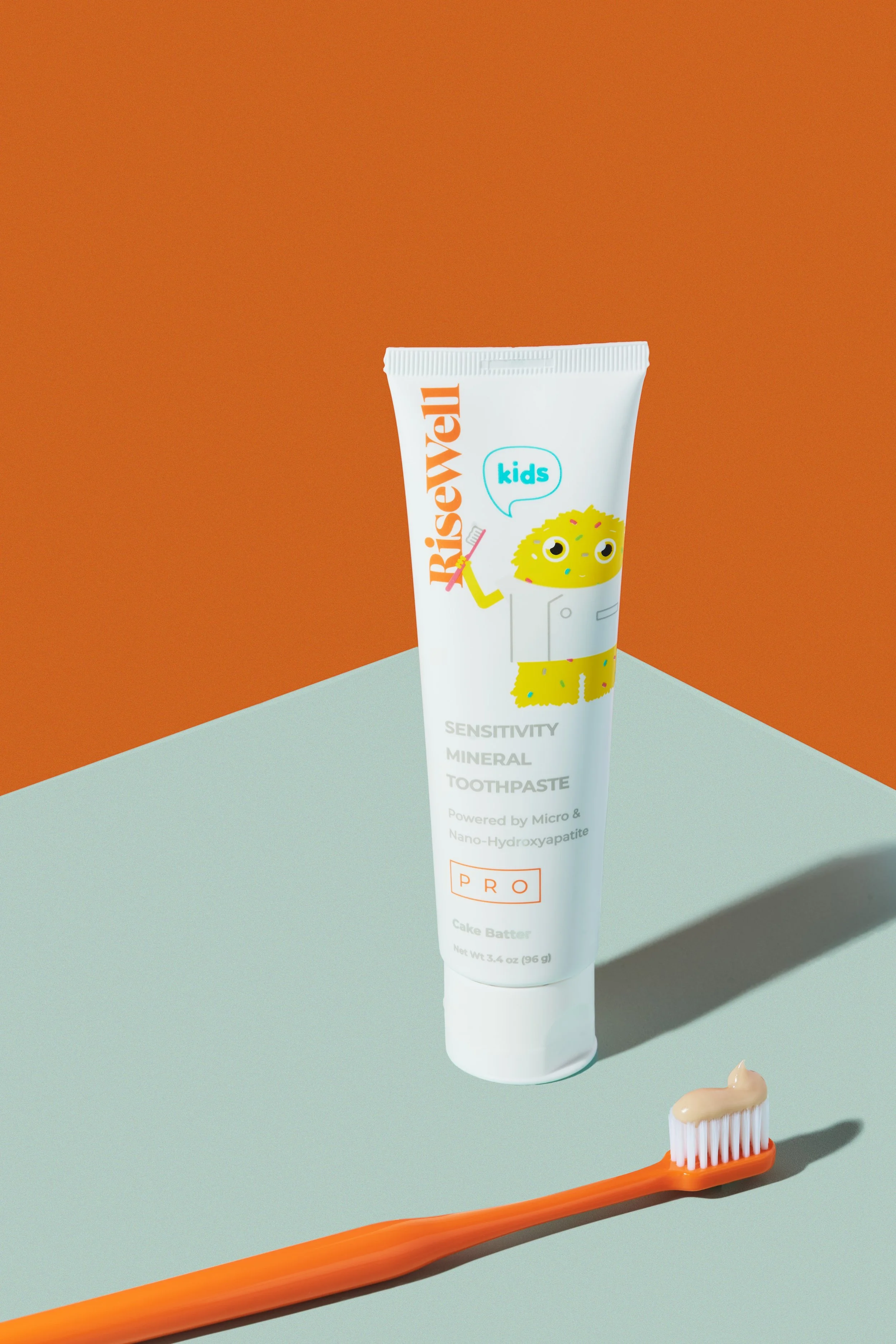 A tube of Risewell kids sensitivity mineral toothpaste with a cartoon monster character, and an orange toothbrush with beige toothpaste on it, set against a dual-colored orange and light green background.