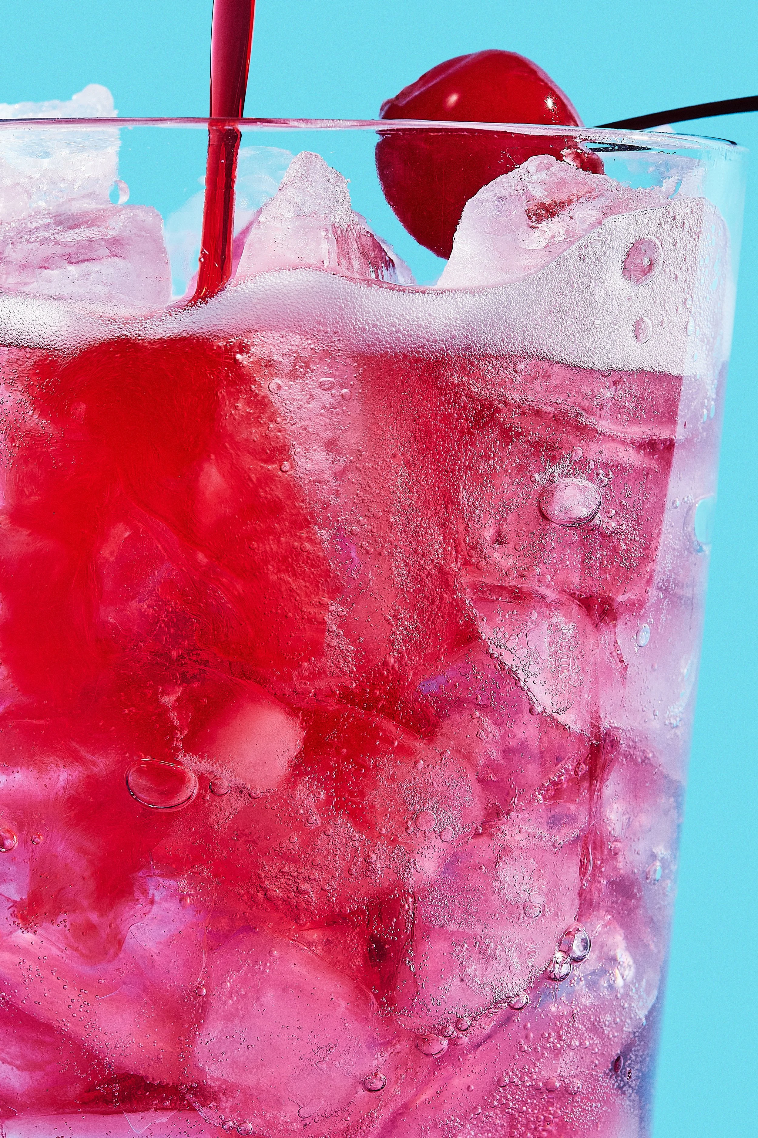 A close-up of a glass filled with a pink beverage, ice cubes, and a cherry on top, with a red straw against a blue background.