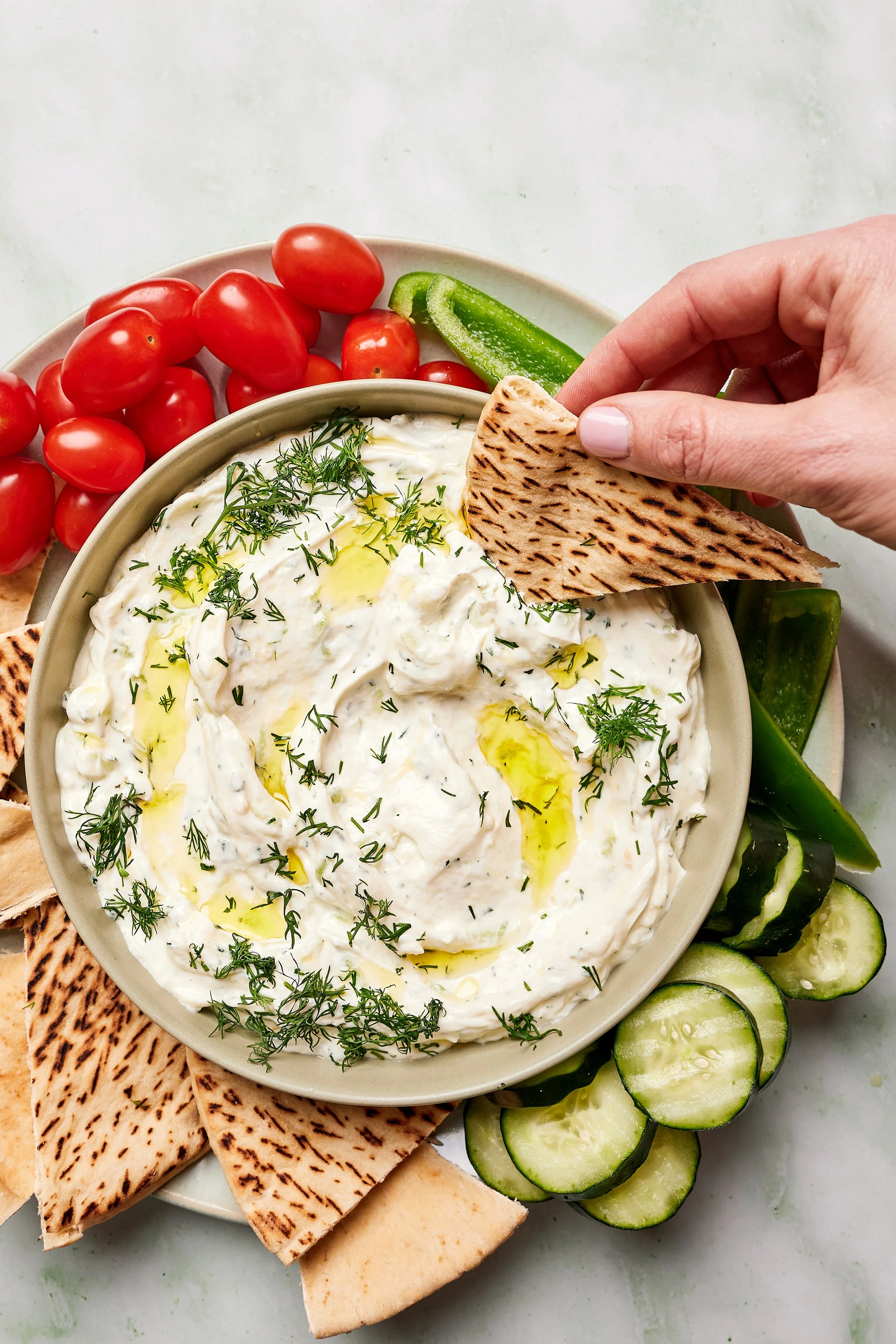 A bowl of creamy yogurt or dip garnished with herbs, with sliced cucumbers, cherry tomatoes, green peppers, and pita bread around it.