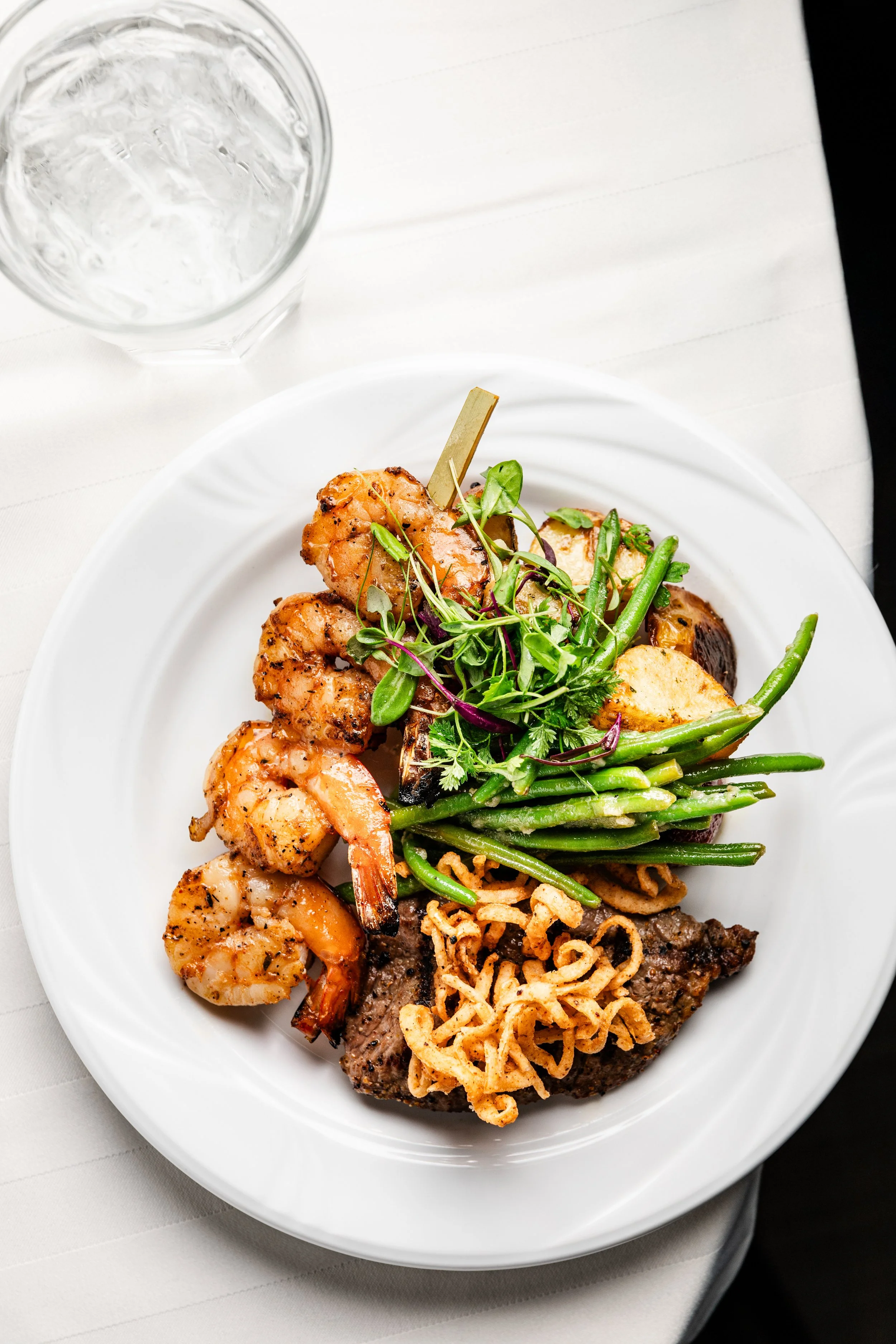 Plate of grilled shrimp, grilled chicken, green beans, salad greens, and fried onion strings, with a glass of water nearby.