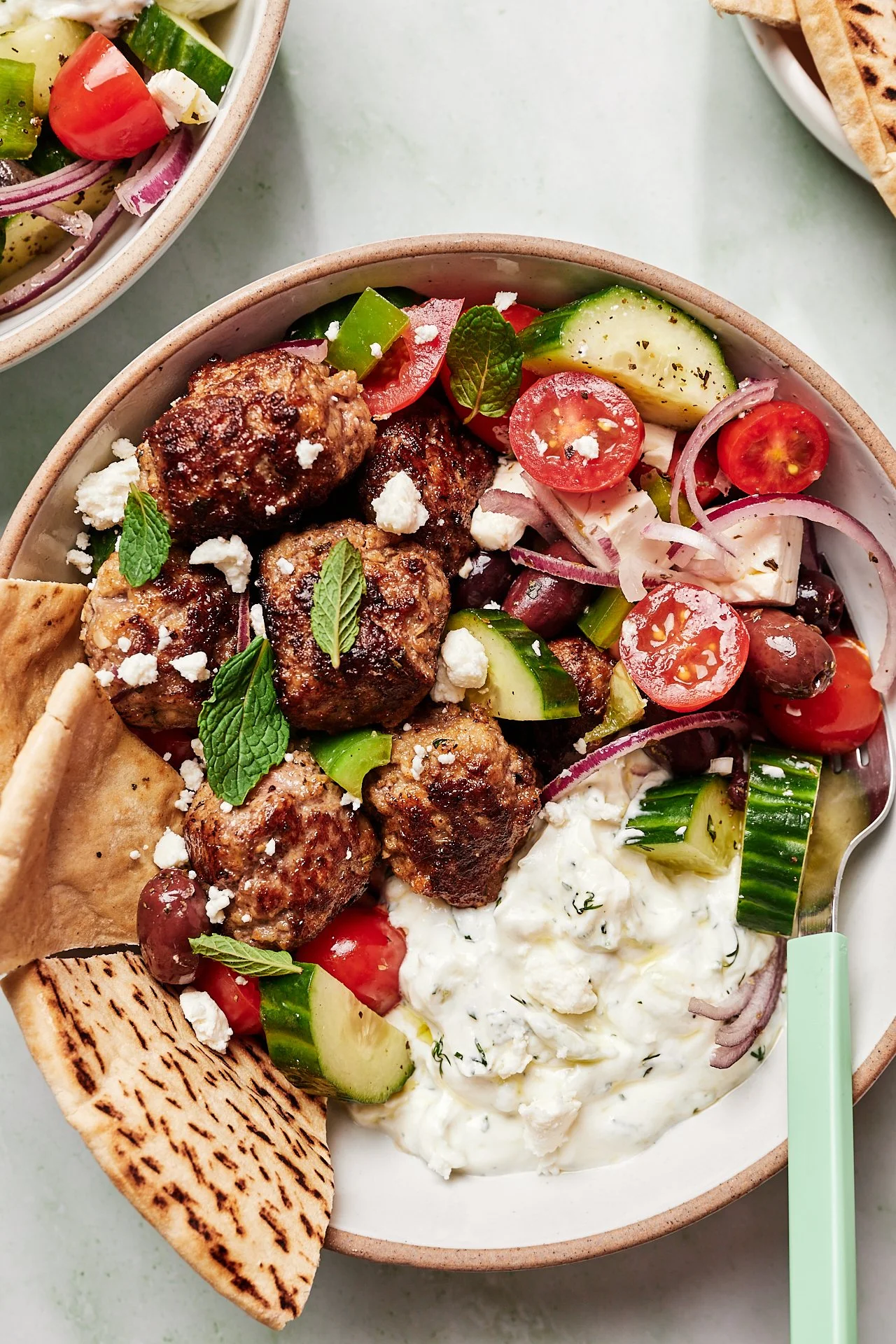 A bowl of Greek salad with meatballs, cucumber, cherry tomatoes, red onions, black olives, feta cheese, fresh herbs, and a dollop of tzatziki, with a piece of pita bread.