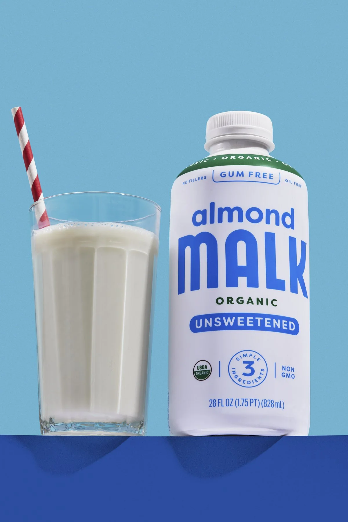 A glass of almond milk with a striped straw next to a carton of almond milk on a blue background.