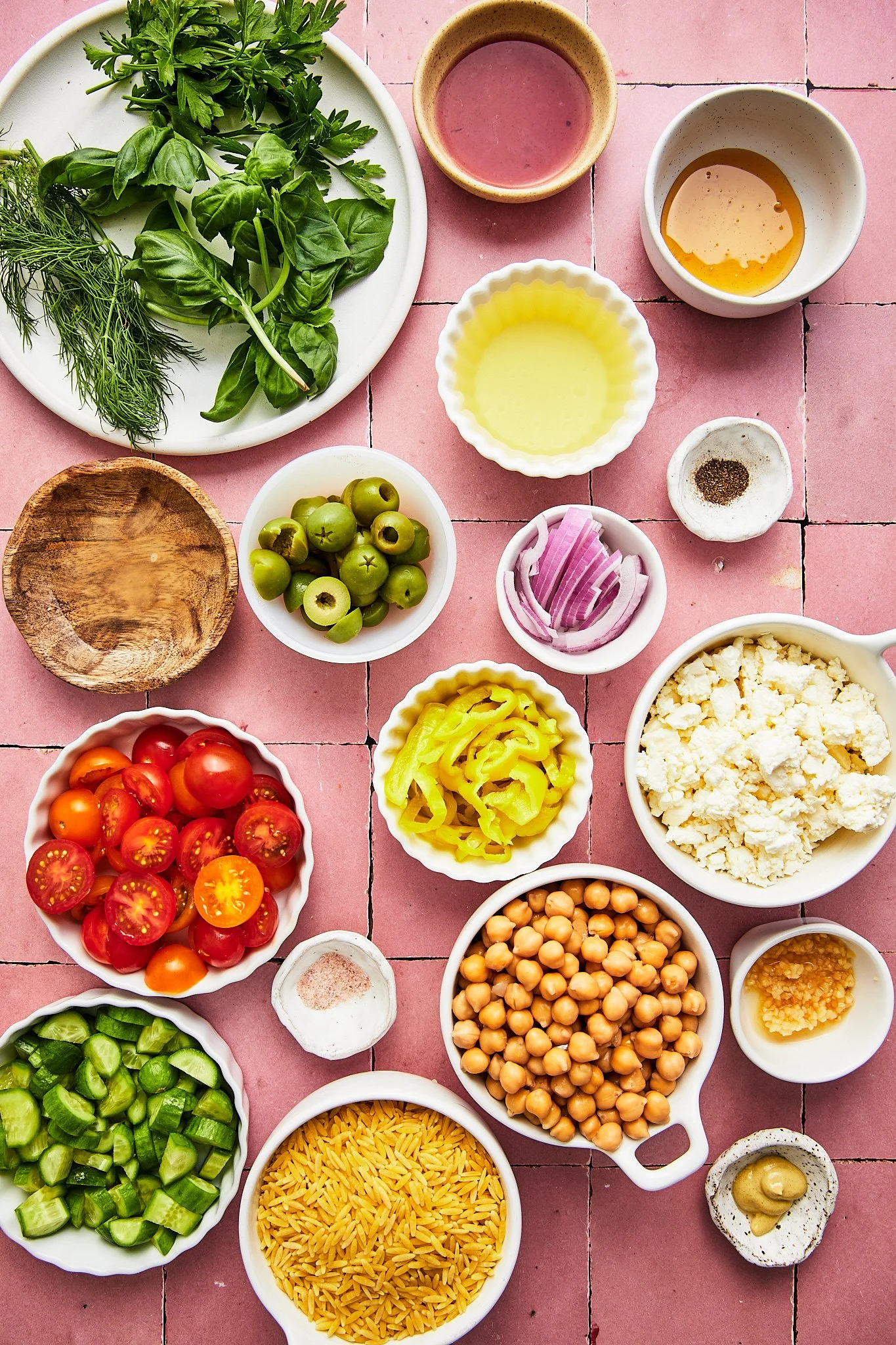 A variety of fresh vegetables, herbs, chickpeas, rice, cheeses, and condiments arranged on a pink tiled surface, likely ingredients for a meal.