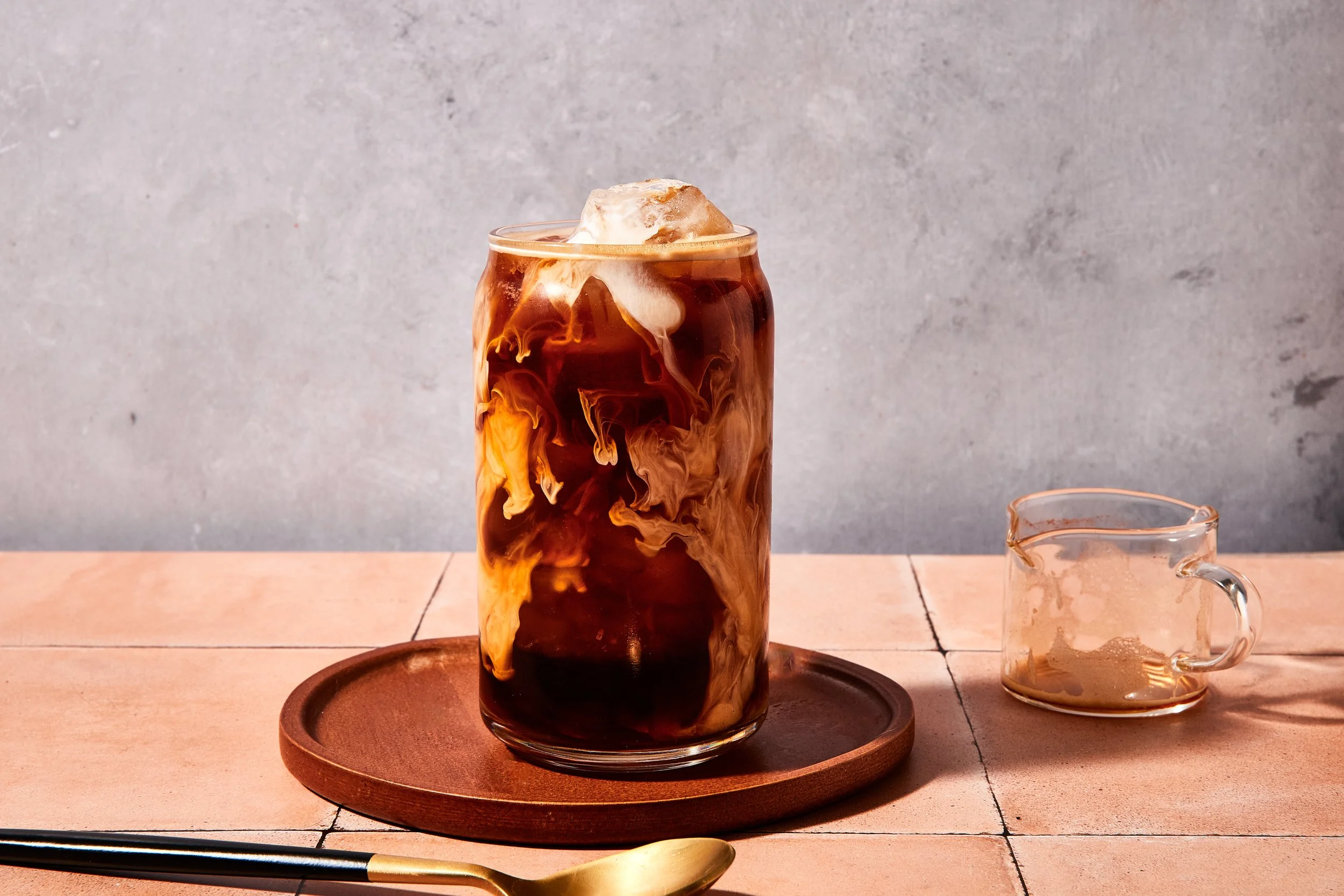 A glass of iced coffee with milk swirls on a wooden tray, placed on a tiled surface next to a small glass creamer and a golden spoon.