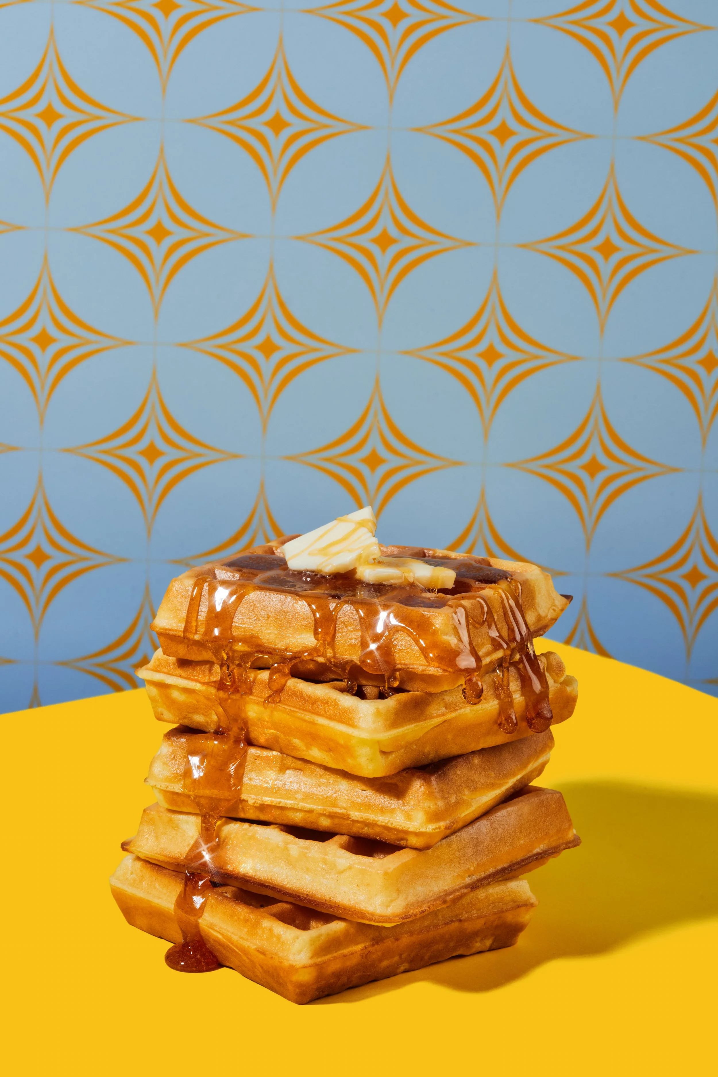 Stack of four waffles topped with butter and caramel syrup, with a patterned blue and yellow background.