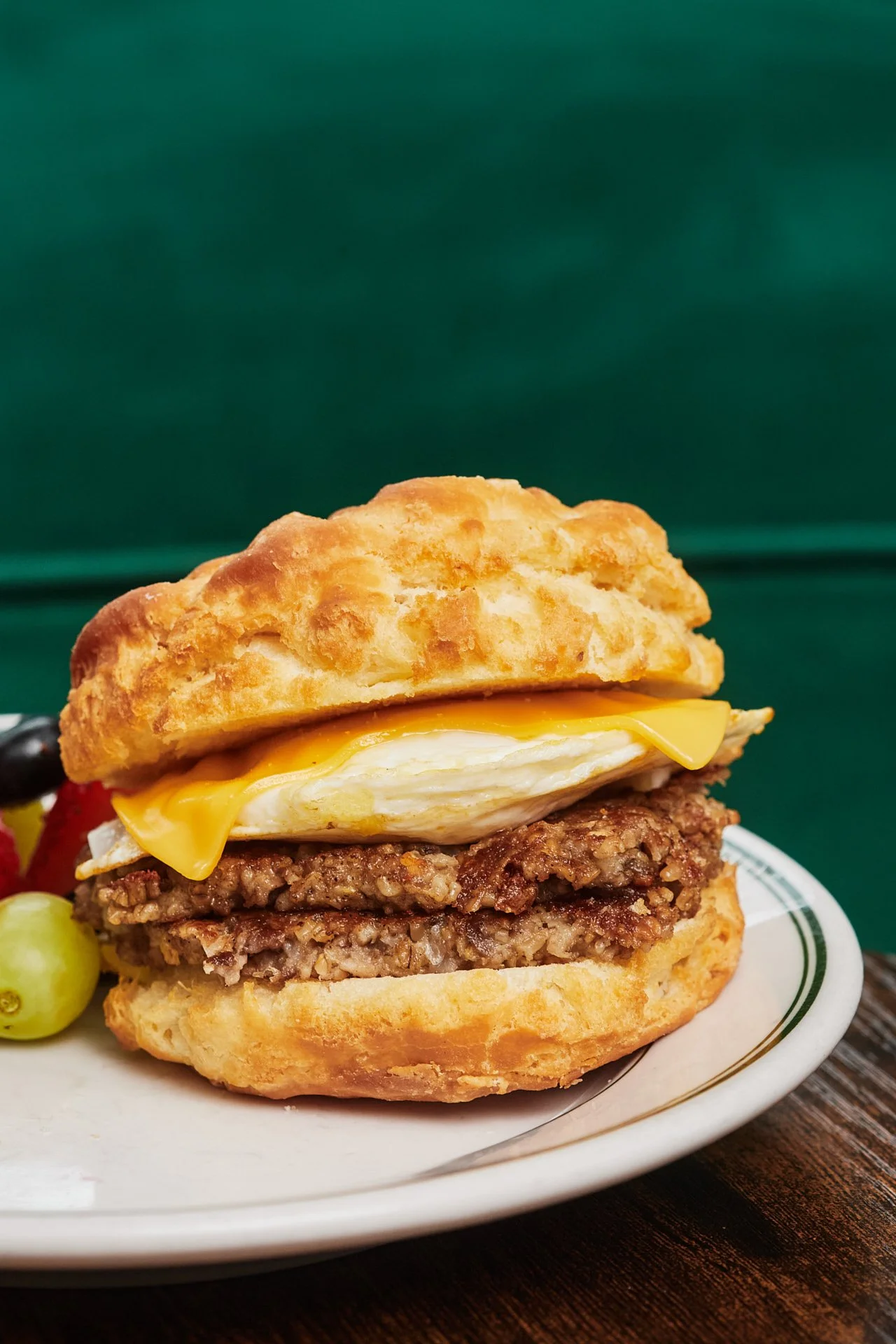 Close-up of a breakfast sandwich with fried chicken patties, a fried egg, and melted yellow cheese on an English muffin, served on a white plate with grapes and berries, against a green background.