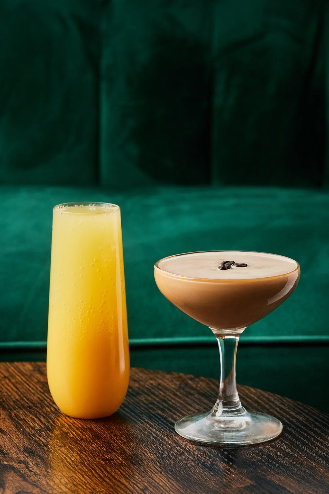 A yellow beverage in a tall, slim glass and a creamy cocktail with coffee beans on top in a coupe glass, placed on a wooden surface.