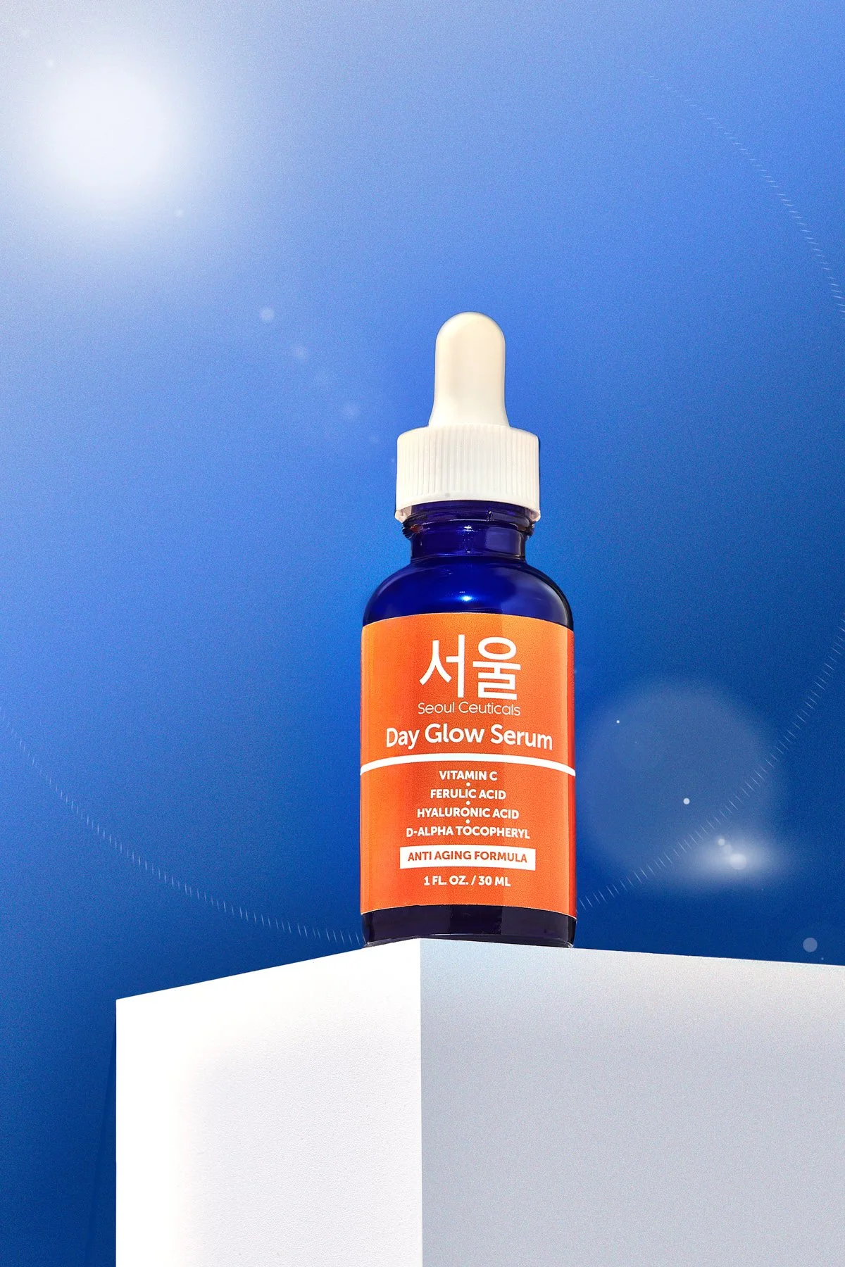 A blue bottle with a white dropper cap labeled 'Seoul Ceuticals Day Glow Serum' on a white platform against a blue background. The label lists ingredients including Vitamin C, Ferulic Acid, Hyaluronic Acid, and D-Alpha Tocopherol, and describes it as