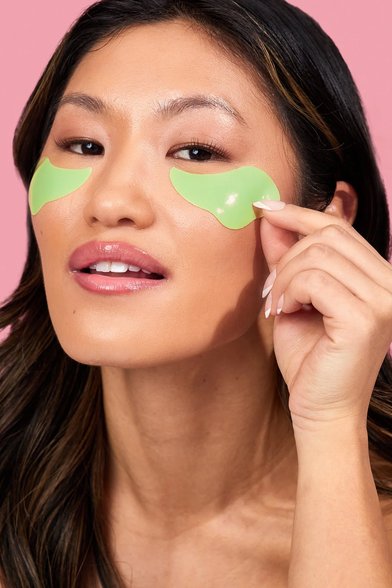 A woman with long dark hair applying green under-eye gel patches against a pink background.