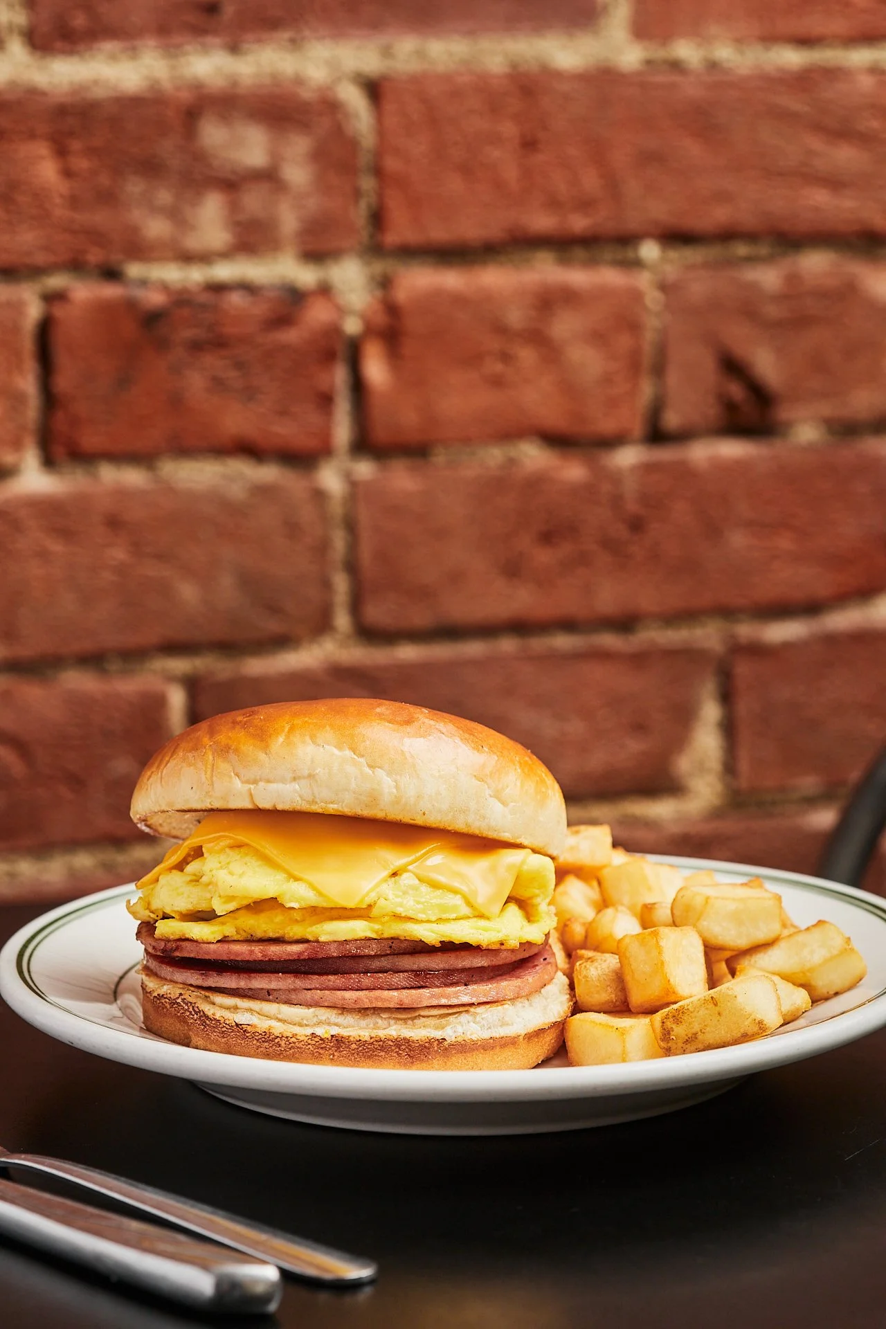 Breakfast plate with an egg and cheese breakfast sandwich with Canadian bacon, and a side of French fries, set against a brick wall background.