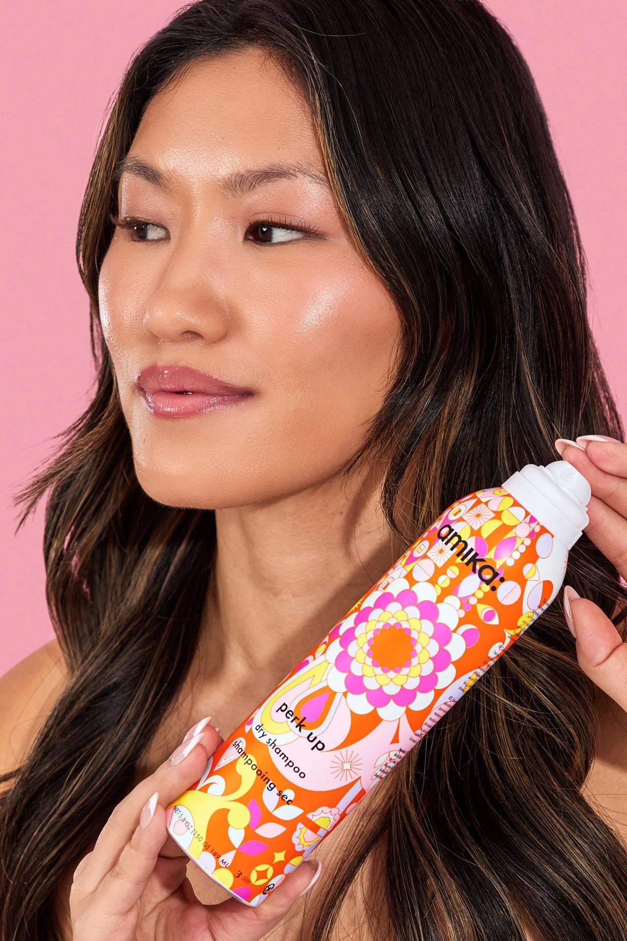 A woman with long, dark hair and a natural makeup look holding an orange and pink floral patterned can of Amika Perk Up dry shampoo against a pink background.