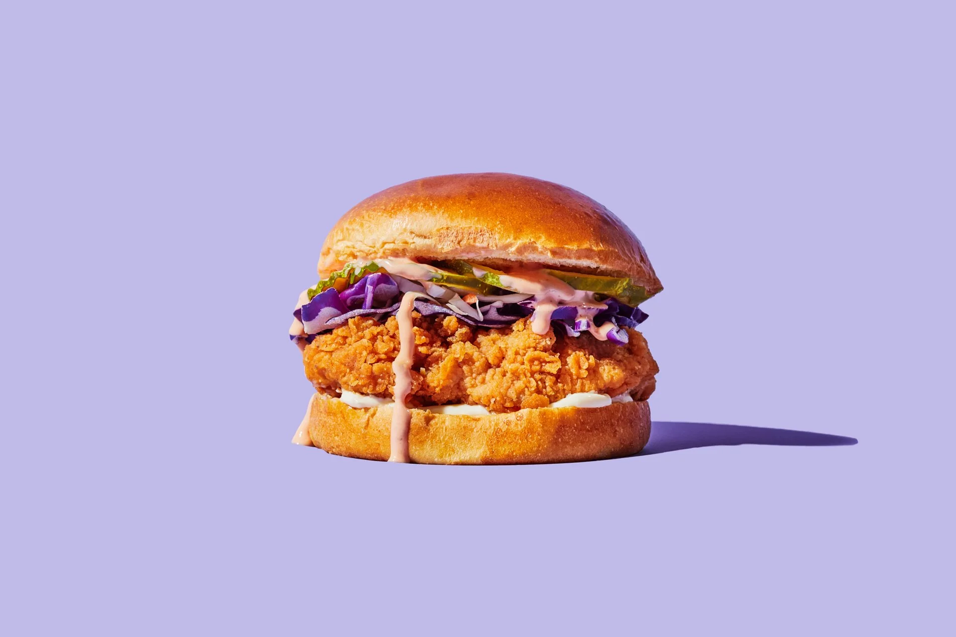 Fried chicken sandwich with purple coleslaw, pickles, and creamy sauce on a brioche bun against a lavender background.