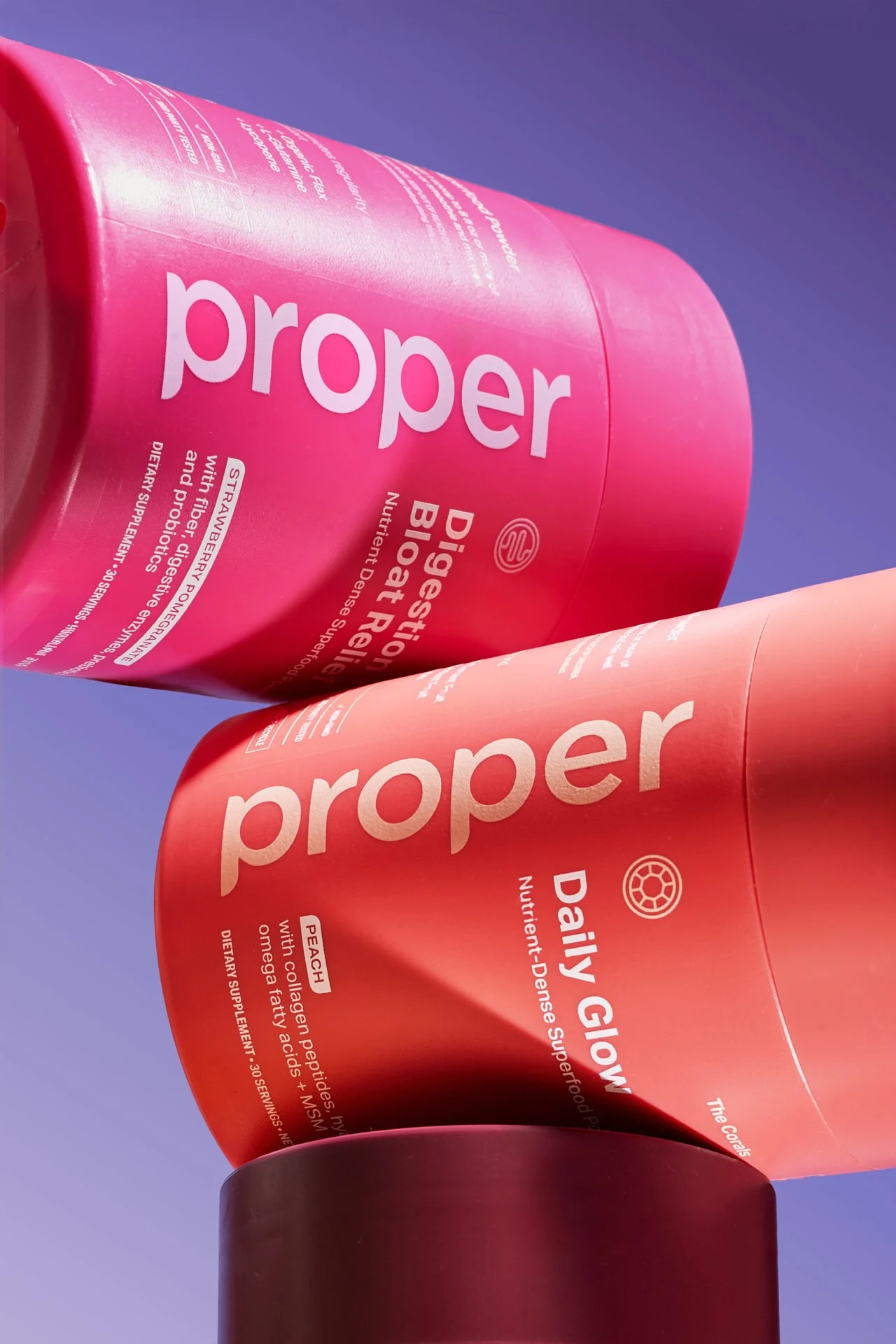 Two cans of Proper brand dietary supplements, one strawberry flavor and one peach flavor, stacked against a blue sky background.