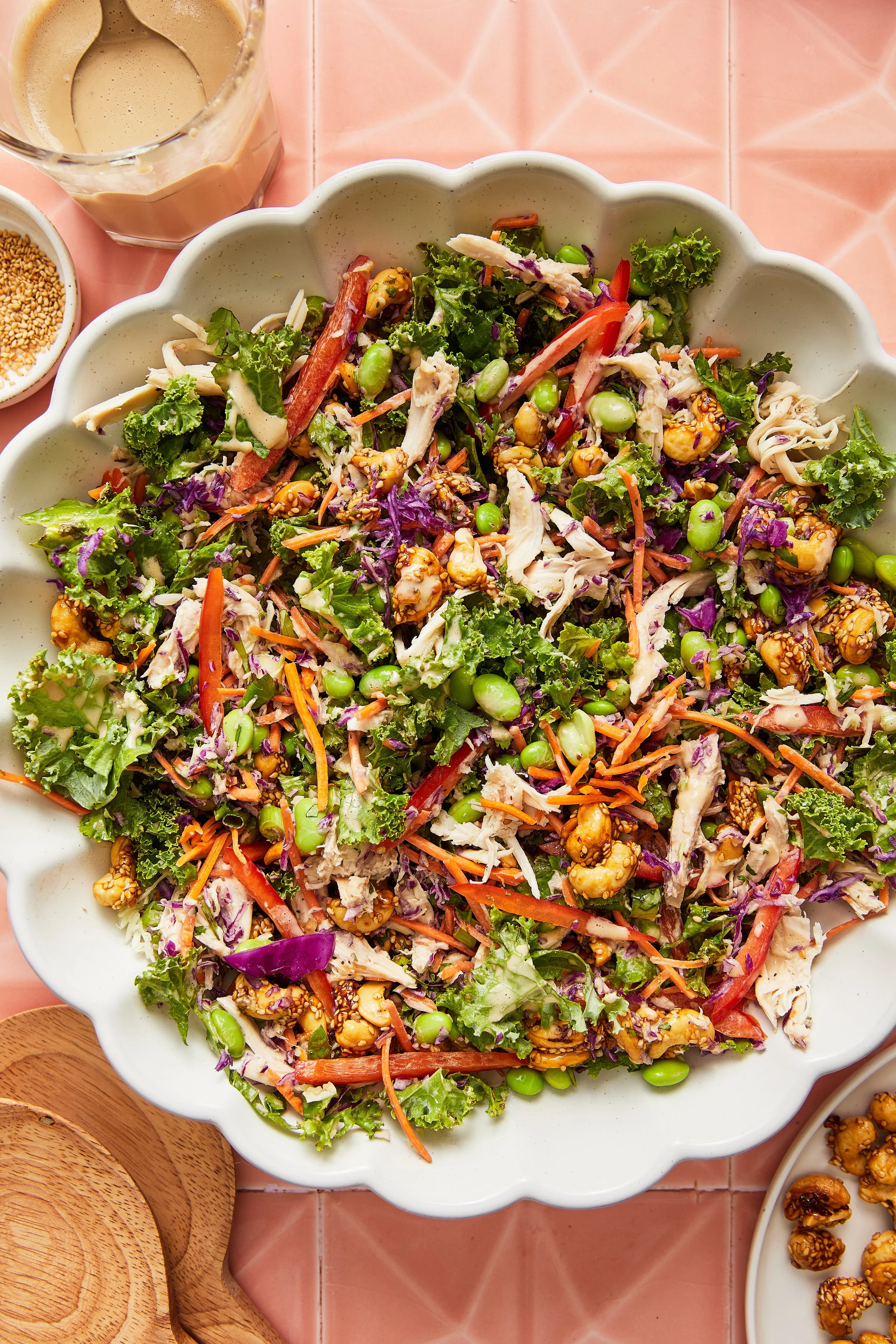 Large bowl of mixed vegetable salad with purple cabbage, carrots, edamame, shredded chicken, and sesame seed-coated nuts on a pink tiled surface.