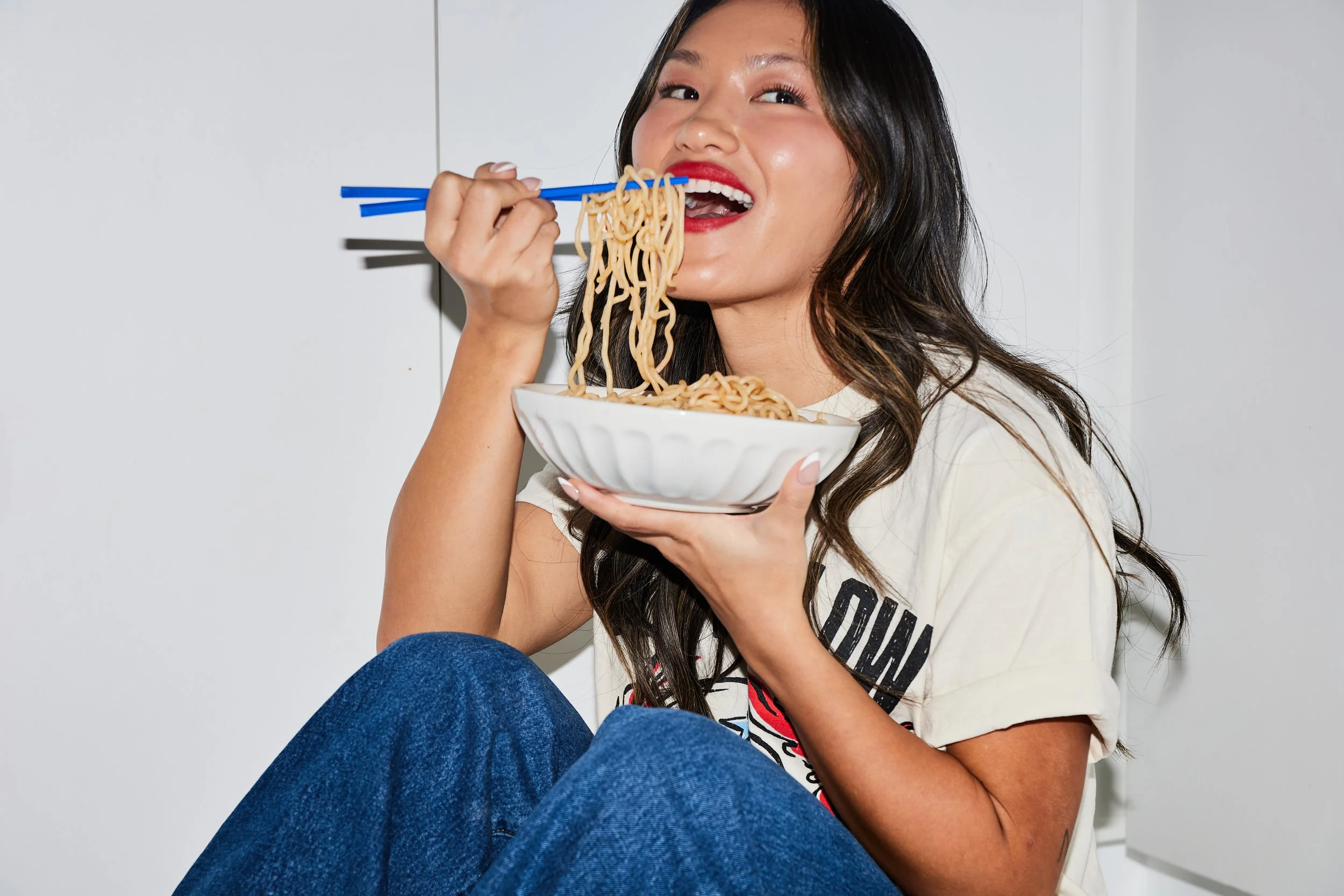 A young woman with long dark hair, wearing a white T-shirt and blue jeans, is smiling and eating noodles with blue chopsticks from a white bowl.