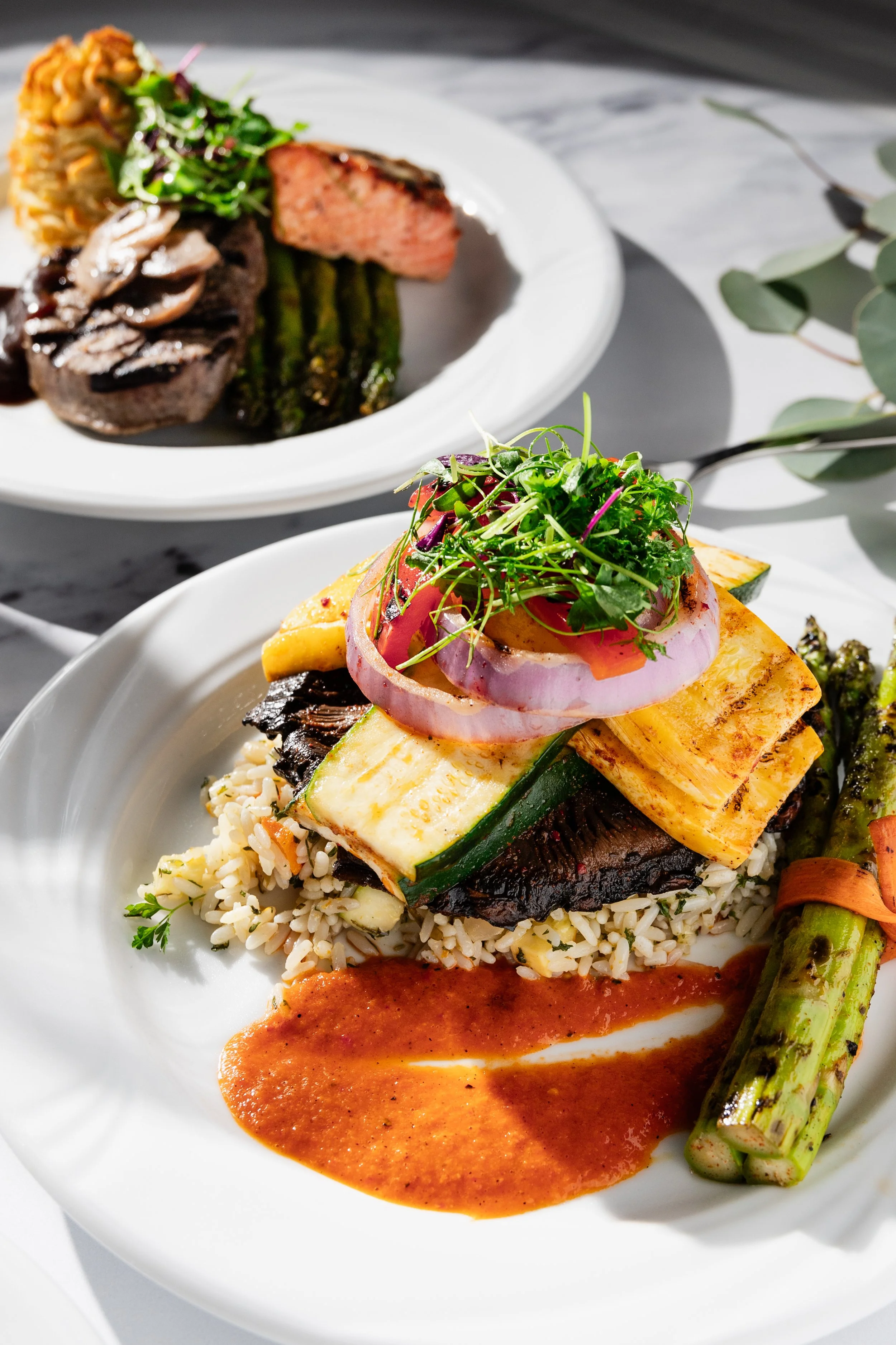 Plated dish with grilled vegetables, rice, and sauce in the foreground, and a plate with steak, mashed potatoes, asparagus, and salad in the background.